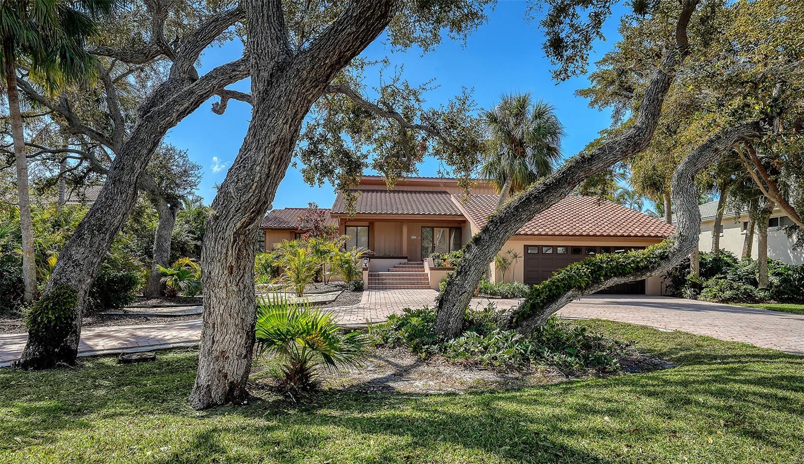 Wrapped in lush, mature landscaping and set along a tranquil mangrove-lined canal, this waterfront 3BR/2.5BA residence on Longboat Key offers space, privacy and an inviting canvas for future vision. A circular paver driveway and stepped front entry create a gracious arrival, framed by tropical greenery and a classic barrel tile roof. Inside, cathedral ceilings with exposed beams, clerestory windows and a wall of glass sliders fill the great room with natural light. A corner fireplace adds warmth and character, while the open layout feels airy and welcoming. Sliders extend the living space outdoors to a wood deck shaded by a pergola, where peaceful canal views set a calming backdrop for everyday living. The kitchen brings a distinctive mid-century influence, featuring low-profile black-and-white cabinetry, a center island with sink, black stone countertops, white appliances, a breakfast bar, an eat-in area and an adjacent dining space that flows easily into the great room. It is functional and full of personality, with room to reimagine if desired. The primary suite is spacious, offering slider access to the deck and a large bath with dual vanities, spa tub and walk-in shower, presenting an opportunity to update and personalize. Guest bedrooms are comfortably sized, and the guest bath is generous with dual vanities. An in-residence laundry room adds convenience. Outdoors, a caged pool with spa sits privately to the side of the home, while the rear of the property opens to excellent boating water. A private dock and boat lift are positioned along a protected canal, bordered by mangroves and just moments from Sarasota Bay. Located in The Bayou, an intimate gated enclave within Bay Isles, residents enjoy access to the private Bay Isles Beach Club, along with optional membership to the Longboat Key Golf & Tennis Country Club and Harbourside Moorings deepwater slips. This is a rare opportunity to enjoy waterfront living today, with the freedom to shape what comes next in one of Longboat Key’s most desirable communities.