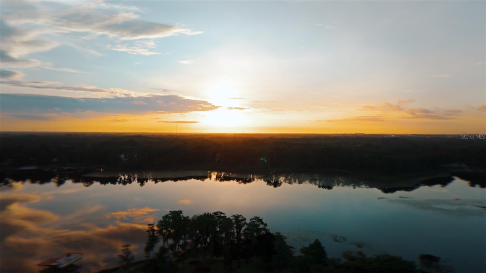 Wake up to a beautiful fire-lit sunrise over the water. On a private 1.6-acre lot with 150 feet of frontage along 83-acre Lake Georgia, a rare ski-friendly lake in East Orlando, 9889 Lake Georgia Drive is an extraordinary custom-built lakefront estate where every detail has been sourced, engineered and installed at the highest level. Offering five bedrooms and six and one-half baths, this is a home defined by scale and craftsmanship. Pass through the sliding gated entry, follow the brick-paved driveway toward the striking custom pivot door, framed in limestone and accented with Turkish stone, and you understand immediately that this is no ordinary address. Inside, the home opens dramatically as 48-by-48-inch porcelain tile floors extend beneath soaring volume ceilings, flanked by floor-to-ceiling windows that draw the lake directly into the living space. Custom floating stairs rise with sculptural elegance beside a crystal chandelier and glass railings, while curated designer lighting sets a tone of refined permanence throughout. A stacked-stone fireplace anchors the great room, and a wet bar with gas fireplace extends the entertaining space. The chef's kitchen is anchored by a large waterfall-edge quartz island, complemented by German custom high-gloss cabinetry, premium stainless steel appliances, a built-in Thermador espresso machine and a porcelain tile backsplash. A walk-in pantry adds everyday function to a home designed to be lived in as beautifully as it looks. The primary suite offers lake views, dual quartz vanities, glass-enclosed rain showers, a handheld bidet and a walk-in closet with custom organizers. Additional en-suite bedrooms feature luxury vinyl plank floors and designer baths with backlit mirrors and matte fixtures. Most rooms are wired for interior and exterior audio, with surround sound in the media and family rooms, all controllable by phone or remote. A dedicated home office, a game/media room ready for billiards, and a second-floor loft complete the interior. Outside, resort-caliber amenities await. A magnificent summer kitchen is outfitted with a 48-inch barbecue, four gas burners, granite counters, ice maker, mini-refrigerator and sink, all beneath a tongue-and-groove wood porch ceiling with automatic screens. The newly resurfaced lap pool features a spa, sun deck, water feature and Pebble Tec surface, surrounded by 2,000 square feet of paver decking and adjoined by a nine-person spa. Above, a balcony finished with artificial turf delivers panoramic lake views. On the water, a large covered boat dock with upper deck, boat hoist, private slip and Sea-Doo storage, plus a private boat ramp on site, completes the picture. Additional highlights include a 22,000-kilowatt generator, dual tankless water heaters, water filtration and softening, $25,000 in motorized custom blinds throughout, an insulated three-car garage with refinished floors and custom storage racks, a security surveillance system, LED lighting, gas-infused double-pane windows and doors, a lake-fed irrigation system and parking for 12-plus vehicles. Ideally located, zoned for Winter Park schools with an Orlando address, just over three miles from UCF and the Central Florida Research Park, home to Lockheed Martin, Northrop Grumman, Raytheon, Boeing and L3Harris. Downtown Orlando and Orlando Health and AdventHealth medical centers are roughly 12 miles west, while Park Avenue's boutiques and dining are minutes away. This is lakefront living at its best.