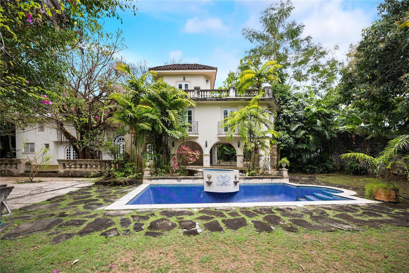 One or more photo(s) has been virtually staged. Majestic Mediterranean Estate in Urb. San Patricio! Discover a timeless architectural gem nestled on 1.5 cuerdas of lush, tree-lined terrain in the exclusive Urb. San Patricio. This Mediterranean-style masterpiece offers an unparalleled blend of elegance, comfort, and natural beauty.
The two-level residence features soaring ceilings in the main living area, accentuated by a striking staircase—an architectural centerpiece that gracefully leads to the upper level. The home is designed with open, flowing spaces perfect for both formal entertaining and relaxed living, including expansive areas for the living and dining rooms.
Outdoors, an expansive open plaza surrounds a captivating Ficus tree, creating a natural gathering point with serene views of the verdant backyard. A covered terrace seamlessly connects to the inviting pool area, ideal for enjoying the Caribbean climate year-round.
Additional features include a detached two-car garage, a utility room, and backup power equipment to ensure convenience and peace of mind. This property also presents an exceptional opportunity for personalization. With remodeling envisioned, it offers the flexibility to incorporate your own needs and desires—whether it’s modern upgrades, custom finishes, or tailored spaces that reflect your lifestyle. We have available architectural drawings and specs for a quick remodeling of the current state. Some photos have been virtually staged for illustration.