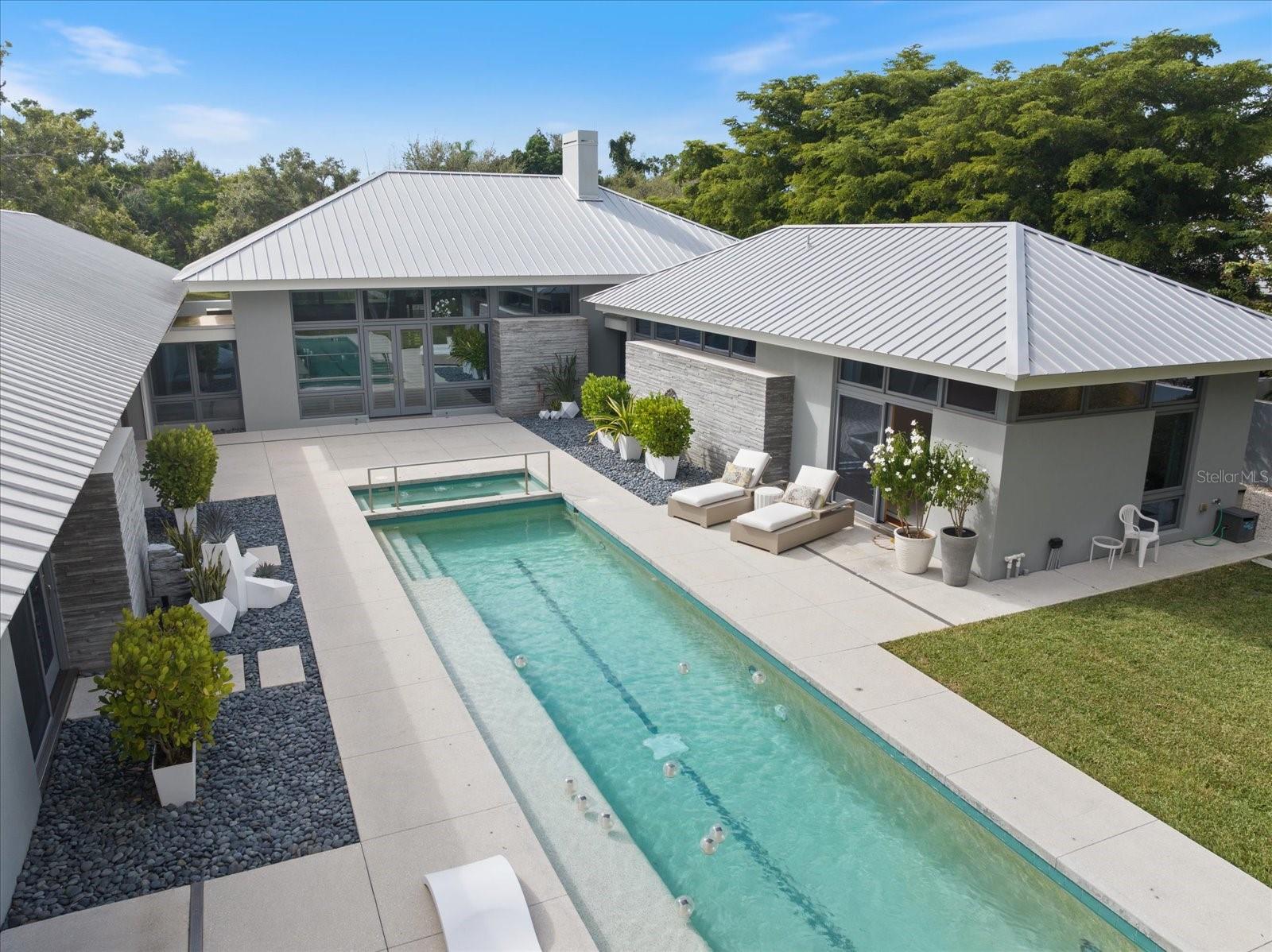 Nestled along Cherokee Drive in the prestigious Cherokee Park neighborhood, this residence enjoys a premier setting on one of Sarasota’s most sought-after streets, renowned for its graceful oak canopy and classic west-of-the-trail charm. This residence embodies the perfect balance of modern architecture, natural materials, and tranquil privacy. Designed by award-winning architect Guy Peterson and constructed by master builder Pat Ball, the home delivers a rare combination of architectural pedigree and enduring craftsmanship.
Positioned on a gracious, estate-sized parcel surrounded by lush greenery, the home’s U-shaped floor plan wraps around its showpiece, a 45-foot crystal-clear pool with integrated spa, creating a seamless indoor–outdoor living experience that defines the Sarasota lifestyle. Outside, the stone pathway leading to the custom mahogany front door sets a striking geometric tone.
Inside, board-on-board architectural molded concrete accent walls, Santos Mahogany wood floors, and cypress-accent ceilings strike a natural harmony with the home’s sleek, modern lines. Floor-to-ceiling impact-rated Marvin windows flood the interior with light, framing tranquil views of the pool and landscape from nearly every room. The wide-open living and dining area, anchored by a wood-burning fireplace, sets the tone for elegant yet effortless entertaining.
The chef’s kitchen features Wolf and Sub-Zero appliances, extensive custom maple cabinetry, and a seamless connection to both indoor and outdoor dining spaces.
In the west wing, the expansive primary suite serves as a private retreat with walls of glass overlooking the grounds, complemented by an adjacent executive office for work-from-home convenience. The east wing hosts two en-suite guest bedrooms, each with sliding glass doors opening directly to the pool.
A detached guest cabana offers a complete living area, kitchenette, and en-suite bedroom that is  ideal for visitors, extended stays, or use as a private art studio or executive home office.
Located within Cherokee Park, one of Sarasota’s most established and beloved neighborhoods, this home offers walking distance to Southside Elementary, minutes to Southside Village and downtown Sarasota, and easy access to Siesta Key Beach, just five streets away. The community is known for its family-friendly gatherings, tree-lined streets, and timeless Sarasota character.