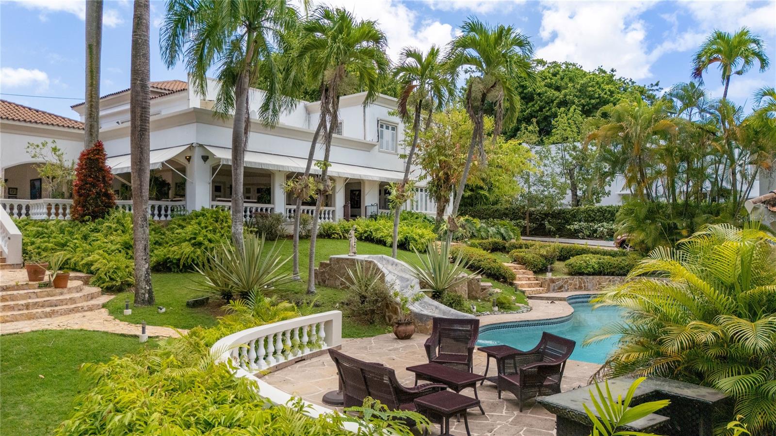 Welcome to one of Villa Caparra's most special homes, ideally located on prestigious A Street, the neighborhood's most desirable address. Set on an extraordinary 2,600-square-meter homesite surrounded by mature trees and lush tropical landscaping, this elegant Spanish Colonial estate offers a level of privacy and serenity that makes it easy to forget you're in the heart of Guaynabo.

Originally built in 1960, the home underwent a thoughtful renovation and expansion in 2000 by a renowned Puerto Rican architect who beautifully preserved its original Spanish character while transforming it into a home designed for today's lifestyle. The result is a residence that feels timeless, welcoming, and effortlessly elegant.

Offering approximately **10,180 square feet of total construction**, including **6,359 square feet of interior living space**, the estate is comprised of a **4,659-square-foot main residence** and a **1,700-square-foot detached guest house and office**.

From the moment you walk through the front door, you're welcomed by soaring ceilings, beautiful red marble floors, graceful archways, and abundant natural light. The formal living and dining rooms open seamlessly onto expansive covered terraces, creating the kind of indoor-outdoor lifestyle Puerto Rico is known for. Whether you're hosting a celebration or simply enjoying a quiet afternoon surrounded by nature, every space feels warm, inviting, and connected to the outdoors.

Upstairs, the main residence offers four spacious bedrooms and three full bathrooms, including a generous primary suite with oversized his-and-hers walk-in closets. At the heart of the home is a chef's kitchen with a large center island that naturally becomes the gathering place, complemented by a temperature-controlled wine cellar just steps away. The main residence also includes two interior powder rooms, a spacious laundry room, and an elevator serving both levels.

The outdoor living spaces are equally inviting, featuring expansive terraces, an outdoor kitchen, two powder rooms, and a full bathroom serving the pool area. Just beyond the pool is a detached two-story guest house featuring a private office with its own full bathroom and storage on the first level, while the upper level offers an independent one-bedroom apartment with a private entrance and full bathroom—ideal for guests, extended family, or live-in staff.

The home is equipped with a 40kW full-power generator, a 10,000-gallon underground water cistern, and a solar water heater, offering comfort and peace of mind year-round.

Homes like this rarely come to market. Rich in character, surrounded by nature, and thoughtfully designed for entertaining, this is a property that offers a lifestyle unlike anything else in Guaynabo.