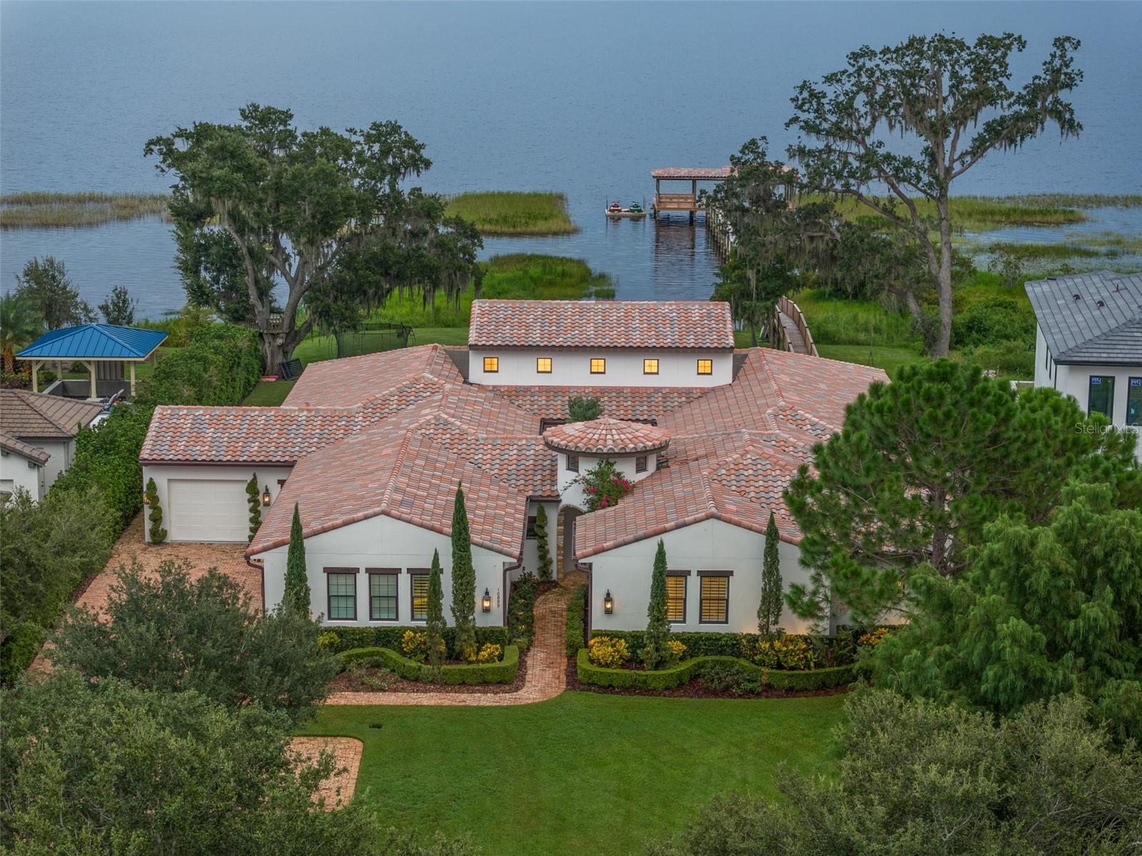 One or more photo(s) has been virtually staged. Welcome home to this stunning single-story, Santa Barbara-style custom residence on the serene shores of Lake Hart, within the gated North Shore golf community. Blending Mediterranean elegance with California rustic charm, the home is surrounded by conservation land and protected state park shoreline, offering both privacy and a true resort-style lifestyle. Built with meticulous attention to detail, this 5,139 sq. ft. energy-efficient home features five bedrooms, four and a half baths, a bonus room, and an interior courtyard. The open floor plan is anchored by a newly renovated gourmet kitchen and great room, each designed to highlight breathtaking lake and pool views. The kitchen showcases premium cabinetry, an oversized island, and a stylish bar area with sink, refrigerator, wine storage, and glass racks—perfect for entertaining.
The primary suite is a private retreat with sweeping lake views, a fireplace, sitting area, dual walk-in closets, and a spa-like bath with a spacious walk-in shower. Additional living spaces include a formal dining room, study, and a bonus room with fireplace that flows seamlessly to the outdoor living area. Step outside to your private oasis, complete with a resort-style pool and spa, outdoor summer kitchen, Sonos surround sound, and upgraded finishes throughout. The backyard also features a covered boat dock with electric hoist, ideal for water sports, fishing, or simply enjoying the sunset over Lake Hart. Unique wood beams sourced from the property add character throughout the home.
Practical luxury continues with a 4-car garage, including one of the garage spaces that has been transformed into an air-conditioned home gym, easily convertible back to vehicle storage if desired. Custom storage throughout the garage provides additional functionality and organization. Conveniently located just minutes from Lake Nona’s Medical City, Boxi Park, the USTA National Campus, top-rated schools, and Orlando International Airport, this home delivers a perfect blend of private resort living with city conveniences. North Shore is anchored by a premier 18-hole championship golf course, elevating the lifestyle in one of Orlando’s most desirable communiti