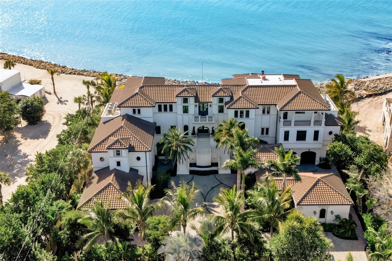 Rising above an enviable stretch of shoreline, this Gulf-front estate showcases timeless architecture and coastal refinement. Designed by CMSA Architects and completed in 2020, the residence features layered rooflines, sculpted stone staircases, and soaring arched windows across more than 180 feet of Gulf frontage. With 10,600 square feet of interiors and 20,982 square feet in total, the home offers a grand yet effortless indoor–outdoor lifestyle, ideal for both elevated entertaining and daily living. A sweeping motor court, mature palms, and a grand double staircase create an immediate sense of arrival. Inside, natural light and uninterrupted Gulf vistas establish a feeling of calm, scale, and sophistication from sunrise to sunset. The great room, lined with floor-to-ceiling arched windows and anchored by a carved-stone fireplace, delivers both serenity and architectural presence. The main kitchen is a triumph of craftsmanship - featuring custom Campbell cabinetry, quartz countertops, designer lighting, Sub-Zero refrigeration, a Wolf six-burner dual-oven range, and a substantial island that anchors the open flow into the living and dining spaces. A prep kitchen and workspace elevate functionality with premium appliances, expansive storage, quartz counters, and a dedicated gift-wrapping wall. The bar and wine room create a refined entertaining destination, defined by custom cabinetry and designer lighting. An iron-and-glass–enclosed, temperature-controlled wine room serves as the focal point, complemented by adjacent illuminated display shelving. Sub-Zero refrigeration, an ice maker, and a Fisher & Paykel dishwasher drawer complete the space. Outdoors, the estate unfolds across three dramatic tiers of Gulf-facing living, featuring arched loggias and a resort-style alfresco kitchen with a wraparound bar that opens seamlessly to the main living spaces. An elevated infinity-edge pool and spa appear to merge with the Gulf, while thoughtfully designed outdoor areas transition effortlessly to a manicured lawn and direct access to the crystal-clear waters at Point of Rocks—where snorkeling, dolphin sightings, and sunset walks become part of daily life. The primary suite forms a private wing, introduced by an illuminated groin-vault corridor that enhances the home’s architectural rhythm. Gulf views fill the bedroom and its terrace, while the bath offers a symmetrically designed retreat with dual vanities, full-height mirrors, multiple chandeliers, a massive double-entry shower, a stone-surround soaking tub, a Sub-Zero–equipped coffee bar, and two exceptional custom closets. Nearby, the office provides a quiet, light-filled workspace. The upper level is devoted to comfort and privacy, offering four well-appointed guest suites, a spacious family room with a wet bar, multiple terraces capturing Gulf views, an additional office, and a laundry room. The ground level is enhanced by indoor–outdoor walkways connecting multiple spaces and generous storage, reinforcing the effortless, resort-style livability. A generator, 3-stop elevator, 6-plus humidity-controlled garage spaces, floor-to-ceiling shades, linear vents, whole-house automation, and shellstone and French oak flooring complete the offering. A short, shaded, tree-lined walk places you at Siesta Key Beach, the No. 1 beach in America by TripAdvisor, offering effortless access to one of the world’s most iconic shorelines.