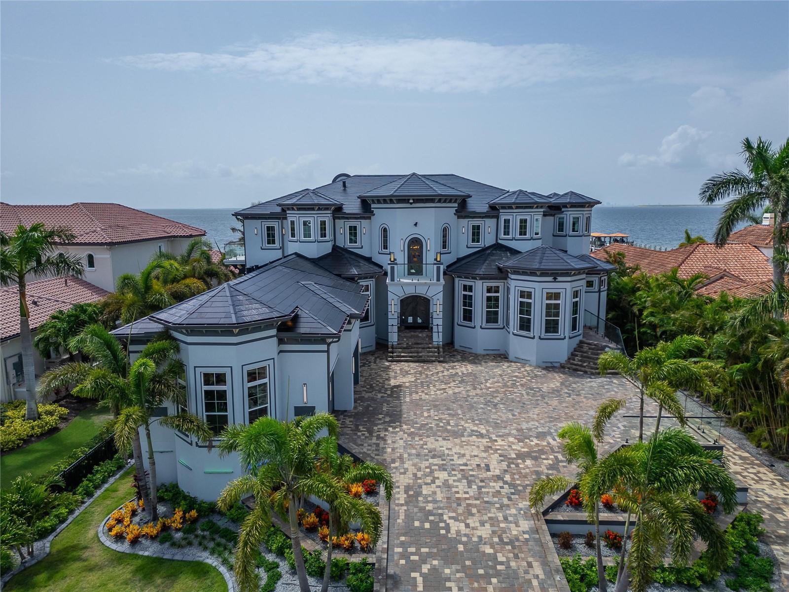 6526 Surfside Boulevard, Apollo Beach FL 33572. 7 bedrooms, 8 bathrooms, 12,896 sq ft heated. Cyprus Signature model by Bistro Builders, completed 2025. Sold fully furnished.
Built in 2025 on 0.58 acres of direct Tampa Bay frontage, this three-story estate delivers panoramic views of the Downtown Tampa skyline and the St. Petersburg waterfront from every level. Tampa International Airport is 30 minutes by car. By water, the covered boat lift dock puts you 25 minutes from Tampa and 25 minutes from St. Pete — a waterfront position unavailable in South Tampa or Davis Islands. The structure is solid block with a concrete floor at every level, stem wall foundation, elevated over 12 feet, hurricane-rated throughout, and fully energy-independent via one of the largest Tesla solar shingle installations in the country, backed by 5 Tesla Powerwall batteries and an LP gas generator.
Inside, an elevator services all three floors. The main level opens to a chef's kitchen with double ovens, gas cooktop, butler's pantry, built-in freezer, wine refrigerator, and a center island scaled for entertaining. The formal dining room is anchored by a backlit custom 200-plus bottle wine room commissioned for this space. An additional 2,000-bottle wine cellar is appointed on the lower level. The primary suite occupies the main floor with private pool access, bay views, a spa bath with jetted tub and walk-in rain shower, dual smart vanities, and a custom dressing room with center island. A dedicated office with separate exterior entrance connects to a safe room and can serve as a security staging area or independent multi-generational suite.
The upper levels deliver a 10-seat custom home theater with French-imported speaker system, a full wet bar and entertainment lounge, wraparound balconies with glass railing and glass floor panels, and a rooftop observatory with unobstructed 360-degree views of the bay and both skylines. Every wall throughout the home is finished with over 10 hand-applied layers of venetian plaster, a master craftsman detail defining the surface quality of each room. Tray ceilings feature commissioned airbrush murals with 24-carat gold inlay by a local artist. A Control4 smart home system manages lighting, climate, security, and audio across all zones. Programmable LED systems cover interior and exterior with infinite configurations.
Garage capacity for 10 vehicles with workshop and gear storage. 5 Tesla charging stations. Whole-home water filtration and softener. Central vacuum. Washer and dryer on both primary floors.
Retractable full corner-pocket sliders on both main levels open the width of the home to the waterfront. The private pool is complete. The outdoor kitchen is built for entertaining when the sliders disappear and the bay becomes the backdrop, featuring a built-in gas BBQ, full grill, dedicated beer tap, outdoor refrigerator, retractable television, and programmable custom lighting.
What the Seller Loves: The rooftop observatory at night. Tampa lit up to the right, St. Pete to the left, the bay between them. The energy independence. The Tesla roof, the 5 batteries, the generator — this home has never lost power and never will. Being 25 minutes from Tampa or St Pete by boat. We are equidistant from both cities and keeping your boat under the covered lift is wonderful. The venetian plaster walls. Every room took weeks. You feel the difference the moment you walk in. This home was built for a principal, not just a family.