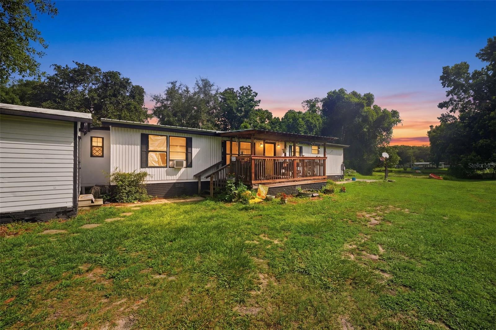 Located on a 0.19-acre corner lot in Floral City, this established 3-bedroom, 2-bath manufactured home with a side-entry 2-car garage offers a comfortable layout surrounded by mature trees. A covered front porch with an elevated wood deck, railings, and steps creates a welcoming entrance and a relaxing place to enjoy the peaceful surroundings. Inside, the spacious living room features laminate flooring that extends throughout much of the home, vaulted ceilings, and large front-facing windows that fill the space with natural light. Adjacent to the living room, the dining area continues the same flooring and provides a bright setting for everyday meals with nearby windows overlooking the outdoors. The kitchen is designed for functionality with extended L-shaped countertops, abundant cabinetry, matching flooring, and windows that brighten the workspace. Just off the dining area, the generously sized family room offers a cozy gathering space centered around a wood-burning fireplace and is filled with natural light from its large windows. The primary bedroom features the same flooring, a built-in closet, windows, and a private en-suite bathroom complete with a single vanity, cabinet storage, a step-in shower, and a window. Bedrooms 2 and 3 continue the same flooring and each include dedicated hanging closet space and windows for natural light. Conveniently located nearby, the guest bathroom features an updated single vanity and a tub-shower combination. The laundry room includes wall-mounted utility shelving for added storage, while the side-entry garage provides additional storage space for tools, equipment, or seasonal items. Outside, the side patio offers a peaceful place to relax while enjoying views of both the front and backyard, all surrounded by mature trees that create a private and tranquil setting. Conveniently located near Withlacoochee State Forest, the Withlacoochee State Trail, and the Tsala Apopka Chain of Lakes, this home provides easy access to boating, fishing, hiking, biking, and the natural beauty that makes Floral City a sought-after destination.