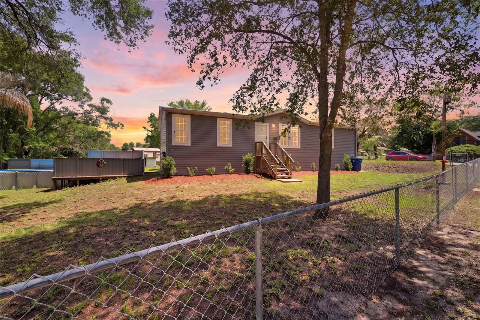 Located on a peaceful 0.38-acre corner lot near the scenic waters of the Withlacoochee River, this 3-bedroom, 2-bath manufactured home offers a unique combination of privacy, versatility, and outdoor enjoyment. Surrounded by lush greenery, the property features a fully fenced yard, an above-ground pool with an adjoining deck, and a detached building complete with its own living area and kitchen space, ideal for guests, hobbies, entertaining, or a private retreat. A front entry staircase welcomes you to the home, where spacious living areas and numerous updates create a comfortable and inviting atmosphere. The open-concept main living areas are designed for comfortable everyday living and effortless entertaining. The spacious living room features plush carpeting and front-facing windows that fill the space with natural light while offering views of the yard. Flowing seamlessly from the living room, the kitchen combines warmth and functionality with luxury vinyl flooring, solid wood cabinetry, solid countertops, stainless steel appliances, a dual sink, and a breakfast bar that serves as a natural gathering spot for casual dining and conversation. The adjoining dining area continues the luxury vinyl flooring and features a backyard-facing window that brightens the space throughout the day, creating a welcoming setting for meals and gatherings. A well-sized primary bedroom offers a relaxing retreat with carpet flooring, a large walk-in closet, and an oversized en-suite bathroom featuring an extended single vanity with solid countertops, abundant solid wood cabinetry, a separate soaking tub with grab bars and solid-surface surround, and a separate step-in shower with sliding doors. On the other side, Bedroom 2 is currently being used as an office and includes carpet flooring and a walk-in closet, while the nearby guest bathroom features a single vanity, solid countertops, solid wood storage cabinetry, and a tub-shower combination. Bedroom 3 offers similar comfort with carpet flooring, walk-in closet space, and a window that allows in natural light. The laundry room is conveniently located near the kitchen and includes luxury vinyl flooring, utility storage, and its own pantry area for additional organization. Outside, an additional staircase provides convenient access from the rear of the home, while the fully fenced backyard provides space to relax and enjoy the outdoors. A raised wooden deck connects directly to the above-ground pool and features privacy walls, creating a tasteful setting for outdoor gatherings, quiet afternoons, or enjoying the peaceful surroundings. Adding even more versatility to the property, an additional building offers its own living area, kitchen space, and double sliding glass doors leading to its own wooden balcony overlooking wooded views, creating a flexible space with endless possibilities. Located in Dade City, this property is near Withlacoochee State Forest, known for hiking, horseback riding, and scenic natural landscapes; Downtown Dade City, offering local dining, antique shops, and year-round community events; and the Suncoast Parkway, providing convenient access toward Tampa, shopping, dining, and travel throughout Central Florida.