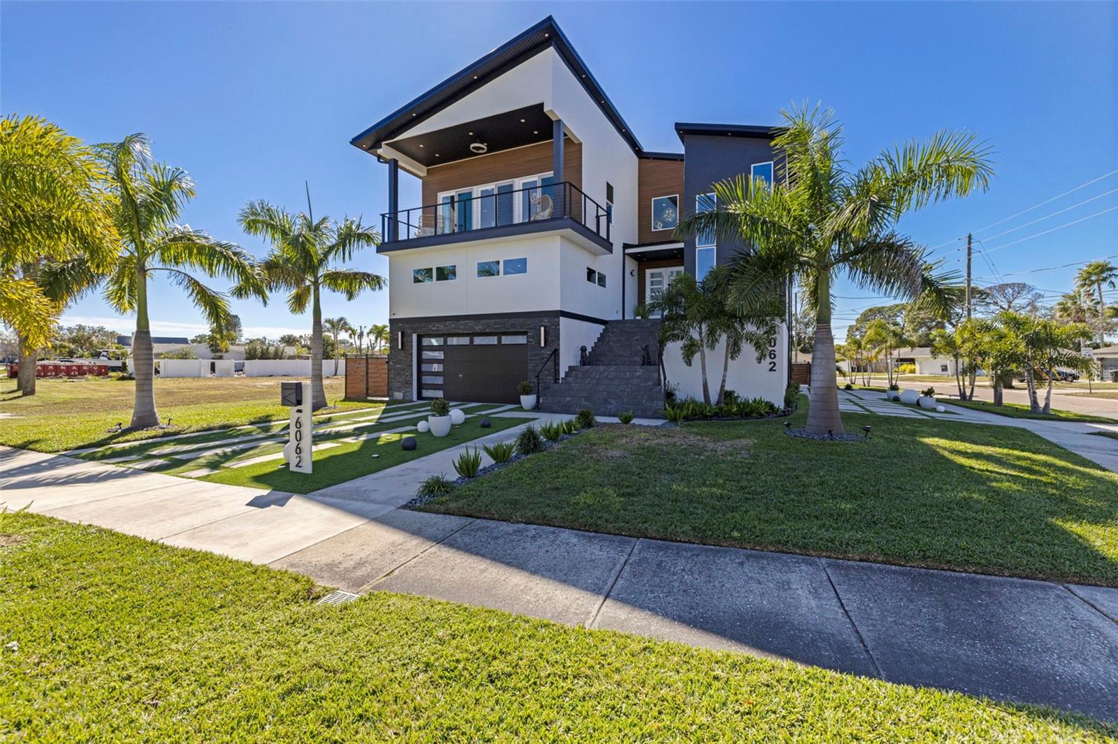 A striking statement of modern luxury, this newly constructed 2023 residence offers over 5,100 square feet of thoughtfully designed living space and more than 9,100 square feet under roof, blending bold architecture, elevated finishes, and resort-style living. Grand front steps lead to dramatic double frosted glass entry doors. Inside, a gallery-style foyer features high-gloss marble-style flooring, layered recessed tray ceilings, and a stunning series of crystal chandeliers that establish the home’s refined, design-forward aesthetic. Thoughtfully layered lighting enhances architectural impact throughout. The main living area impresses with soaring ceilings, expansive walls of glass, and an open mezzanine overlook, creating a bright, connected living experience with dramatic vertical volume. The kitchen is designed for both everyday living and entertaining, anchored by a massive waterfall-edge island and complemented by sleek cabinetry, wall oven, cooktop, pot filler, and two built-in beverage refrigerators. A substantial walk-in pantry with custom shelving adds exceptional storage, while the space flows seamlessly into the dining and living areas. This five-bedroom, six-bath home offers a highly functional layout. Four bedrooms are upstairs, including a private guest suite with en suite bath. One additional bedroom with full bath is located on the main level. A dedicated pool bath, upstairs family room overlooking the main living area, and two laundry rooms, one upstairs and one downstairs, enhance everyday convenience. The primary suite is a private retreat featuring a statement accent wall, custom cut tray ceiling, and designer chandelier. The spa-inspired bath includes a massive walk-in shower with multiple showerheads and rain head, freestanding soaking tub, and a single vanity with dual sinks and an illuminated mirror. A glass-enclosed boutique-style display closet, additional oversized walk-in closet, and private balcony with electric blinds and water views complete the suite. Outdoor living rivals a private resort, with expansive terraces and patios finished in premium honed natural marble. The pool features a sun shelf and integrated spa and is complemented by a fully equipped outdoor kitchen and multiple lounge and dining areas ideal for entertaining. The exceptionally expansive garage is fully finished with polished epoxy floors, recessed lighting, and soaring ceilings, offering space for high-end vehicle collections, a golf cart, or recreational toys, with capacity for approximately ten vehicles. A game or lounge setup with pool table and an interior overlook from the main level elevate the space beyond a traditional garage. The home is fully hurricane rated with impact windows and impact-rated doors throughout. Storage is abundant, from custom closets and built-ins to pantry and garage capacity. This bold modern residence delivers scale, design, and craftsmanship in one exceptional offering. Ideally located within a sought-after waterfront community in the desirable Northeast area, the home is surrounded by scenic waterways, neighborhood parks, and playgrounds, offering an exceptional balance of luxury living and everyday convenience. Residents enjoy walkable green spaces, water access, and a golf cart–friendly lifestyle, with easy access to Islander Market, along with access to A-rated Shore Acres Elementary School and just minutes from vibrant downtown dining and shopping.