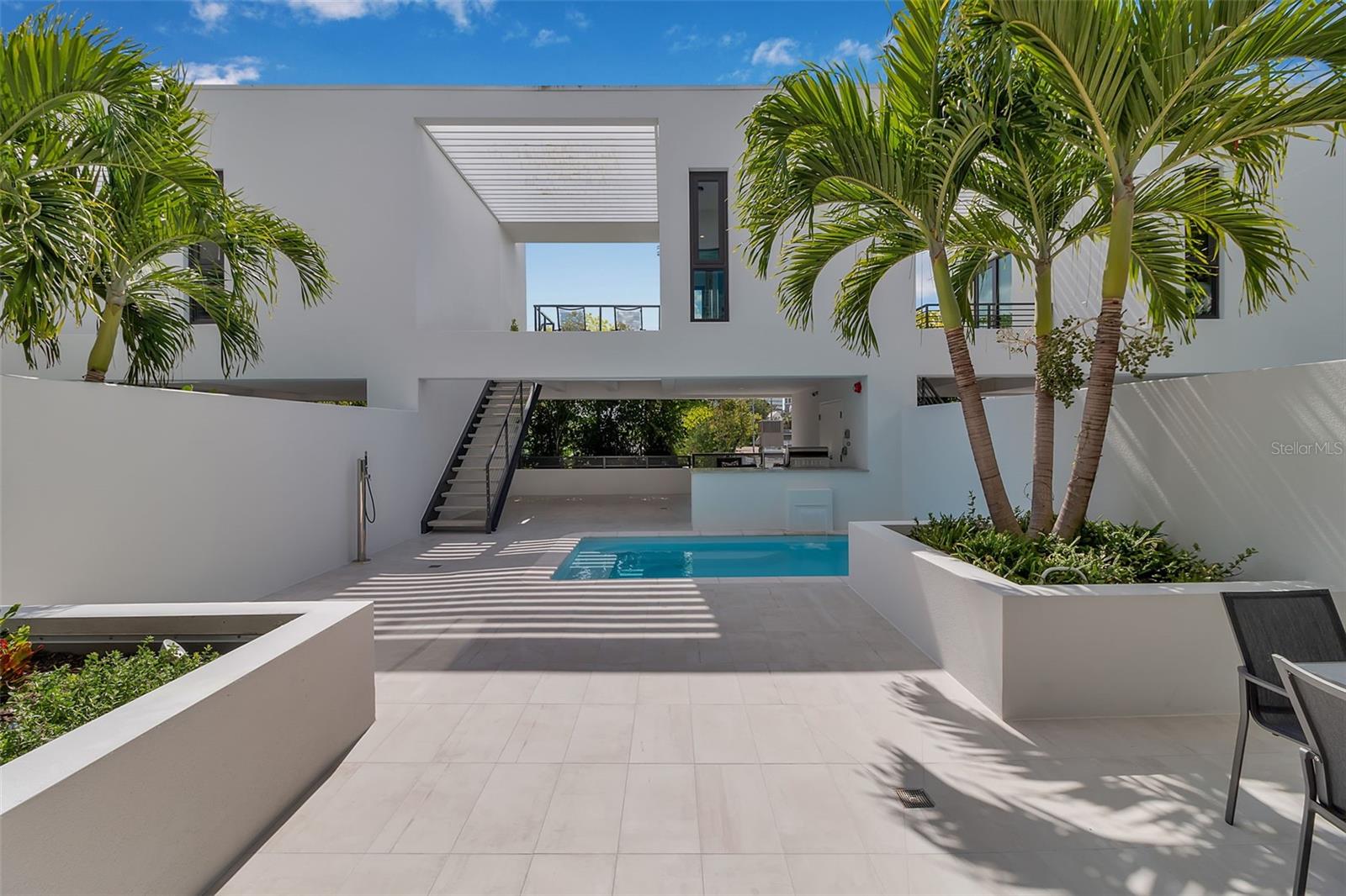 Anchored in Sarasota’s vibrant Rosemary District, a hub of art, dining, and design just steps from downtown, this contemporary multi-level residence captures the essence of urban coastal living. Defined by its clean architectural lines, striking white façade, and geometric precision, it offers a rare balance of privacy and proximity in one of Sarasota’s most walkable neighborhoods. A sleek iron-and-glass entry framed by textured tile sets the tone for what’s inside: light, volume, and effortless connection to the outdoors. The floating staircase, with open risers and maple treads, becomes a sculptural centerpiece beneath ceilings that seem to soar forever.  A three-stop elevator ensures convenience from the ground-level garage to the uppermost terrace. The main living level unfolds with gallery-like openness, a seamless blend of style and function. The sleek kitchen, featuring quartz countertops, premium stainless-steel appliances, and clean-lined cabinetry, overlooks the dining and great room. The open living space is complete with expansive sliders that open directly to the courtyard and its tranquil Zen pool, where the rhythm of water and greenery softens the modern form. With its summer kitchen, fire table, and covered dining terrace, the home transitions effortlessly from quiet mornings to lively evenings. Upstairs, the primary suite feels like a retreat, complete with a glass-framed balcony, spa-inspired bath, and thoughtful details that invite calm. Two additional guest rooms, one that is currently used as an office, mirror the home’s minimalist aesthetic and abundant natural light. A private guest casita, accessible from the courtyard, expands the home’s flexibility. With a kitchenette, full bath, and its own balcony, it’s ideal for visitors, as another primary suite or as a fitness space, all with the independence of a separate entry. Constructed of concrete and steel with impact-rated glass, every element under scores quality and low-maintenance design, perfect for those seeking the ease of a lock-and-leave lifestyle. Positioned in Sarasota’s coveted Rosemary District, this residence places the best of the city just outside your door. Stroll to downtown dining, art galleries, live music, and cultural venues, or stop by the weekly Sarasota Farmers Market. Boutique shops, theaters, and cafés line the nearby streets, while the beaches, the John Ringling Bridge, St. Armand’s Circle, and Sarasota Bay are only minutes away. With convenient access to Sarasota Bradenton International Airport, this address blends logistics, urban luxury, outdoor living, and strong architectural design, offering a truly unique downtown lifestyle.