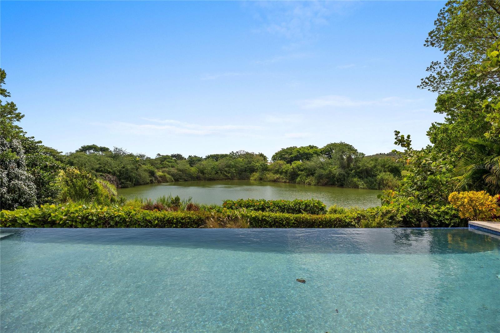 Nestled within the highly sought-after gated community of Sabanera Dorado, this exceptional Carbia model sits on one of the neighborhood's most desirable lots, offering a rare combination of privacy, lake views, generous outdoor space, and thoughtfully upgraded interiors.

From the moment you enter the home, your attention is drawn to an oversized picture window that perfectly frames the infinity-edge pool and tranquil lake beyond. The dramatic entry, complemented by high ceilings and the Carbia model's signature mezzanine design, creates an immediate sense of openness and elegance that distinguishes this home from others in the community. 

The main living areas flow seamlessly into the outdoor spaces, where a wraparound covered terrace invites year-round enjoyment. Surrounded by lush landscaping and peaceful water views, the terrace evokes the charm of country living while offering all the conveniences and amenities of Sabanera Dorado.

Unlike many view lots, this property also features a substantial yard, providing ample space for recreation, entertaining, and outdoor enjoyment. At the heart of the home, the kitchen opens effortlessly to the family room and den, creating a warm and functional gathering space. An expanded pantry offers valuable additional storage and enhances everyday convenience.

The primary suite occupies its own private wing and serves as a luxurious retreat. Featuring vaulted ceilings, additional storage space, and a custom enlarged walk-in closet, the suite combines comfort with functionality. The recently remodeled spa-inspired bathroom includes dual vanities, an oversized walk-in shower, and a beautiful clawfoot soaking tub designed for relaxation outside. Three additional bedrooms are generously sized and each feature their own en-suite bathroom, providing privacy and comfort for family members and guests.

A spacious laundry room adds further practicality to the home's thoughtful design. One of the home's most unique features is the upstairs guest suite. Unlike many mezzanine spaces found in similar models, this residence offers a true private bedroom with its own en-suite bathroom. The suite enjoys both privacy and connection, with an interior overlook that captures views of the living spaces, pool, and lake below.

Combining architectural character, stunning views, expansive outdoor living, and one of the best lots in Sabanera Dorado, this is a rare opportunity to own a truly special home in one of Puerto Rico's premier communities. The property includes a 15KW Diesal Generator, 300 Gallon Cistern and water softener "RainSoft" system. 

Residents of Sabanera Dorado enjoy access to exceptional amenities, including scenic golf cart trails, a fitness center, lap pool, convenience store, and a full-service restaurant. Families especially value the presence of TASIS Dorado, the school founded in partnership with The American School in Switzerland.