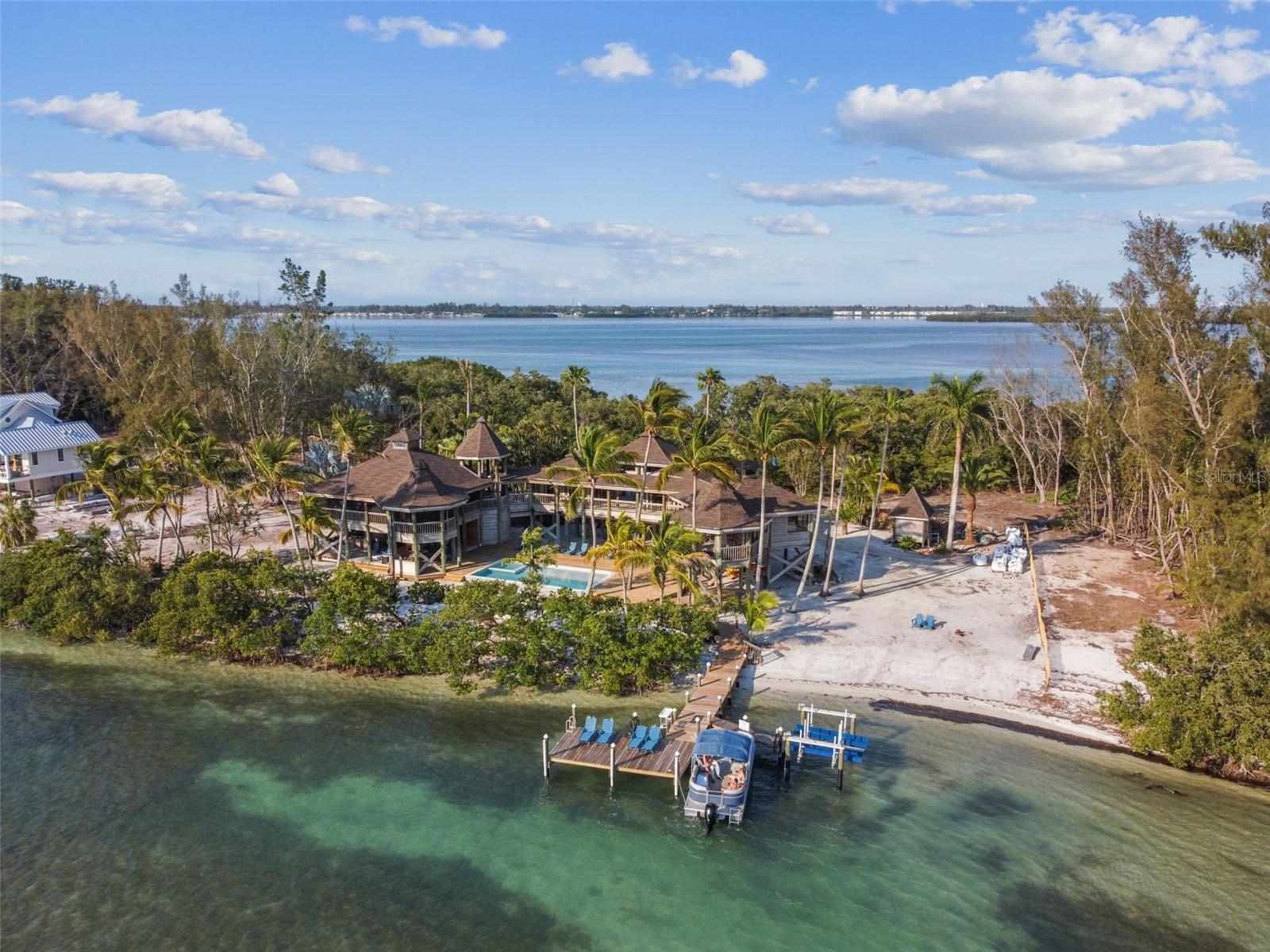 A rare offering on Florida’s coveted west coast, this private island sanctuary feels worlds away yet remains moments from the region’s most beloved coastal destinations. Set on the exclusive 38-acre island of Jewfish Key (accessible only by boat), this remarkable 1.35-acre estate commands its own secluded shoreline and uninterrupted views of crystal clear waters. Inspired by Polynesian architecture and designed to dissolve the boundaries between indoors and out, this estate home unfolds through airy pavilions connected by elevated walkways. Towering palms and lush tropical landscaping envelop the home, while its 18-foot elevation ensures both peace of mind and panoramic, horizon-level vistas. Inside, the freshly reimagined kitchen serves as the gathering heart of the home, with sleek quartz countertops, modern cabinetry, and a spacious oversized island that effortlessly bridges culinary function and relaxed entertaining. Expansive sliding glass doors draw natural light into the living space and frame sweeping views of the sandbar and bay, perfect for sunset watching. The main residence includes four bedrooms and four-and-a-half baths, plus a separate guest suite offering an additional bedroom, kitchen, and full bath, ideal for hosting with comfort and privacy. The primary suite is where the pace of the island truly settles in. Wake up to wide-open bay views, linger with coffee in the sitting area, or enjoy a glass of wine as the sun sinks toward the horizon. The ensuite feels fresh and spa-like, with a soaking tub, walk-in shower, and water views. A series of screened porches and wraparound decks encourage slow living, morning yoga on the deck, afternoon reading in the shade, sunset grilling and cocktails overlooking the Gulf. Outdoors, a newly installed pool and spa with a sun shelf and expansive sundeck overlook the storied Jewfish Key Sandbar. A private dock with jet ski lift offers instant access to calm waters perfect for fishing, snorkeling, paddle boarding, or exploring neighboring Longboat Key and Anna Maria Island, both just minutes away by boat and beloved for dining, boutique shopping, and laid-back coastal culture. Equal parts island hideaway and refined coastal home, this one-of-a-kind property offers the rare combination of true privacy, effortless access, and natural beauty, set against a backdrop of turquoise water, powder soft sand, and endless sky. Here, relaxation, adventure, and natural beauty converge in a setting that feels almost like an escape, yet unmistakably home.