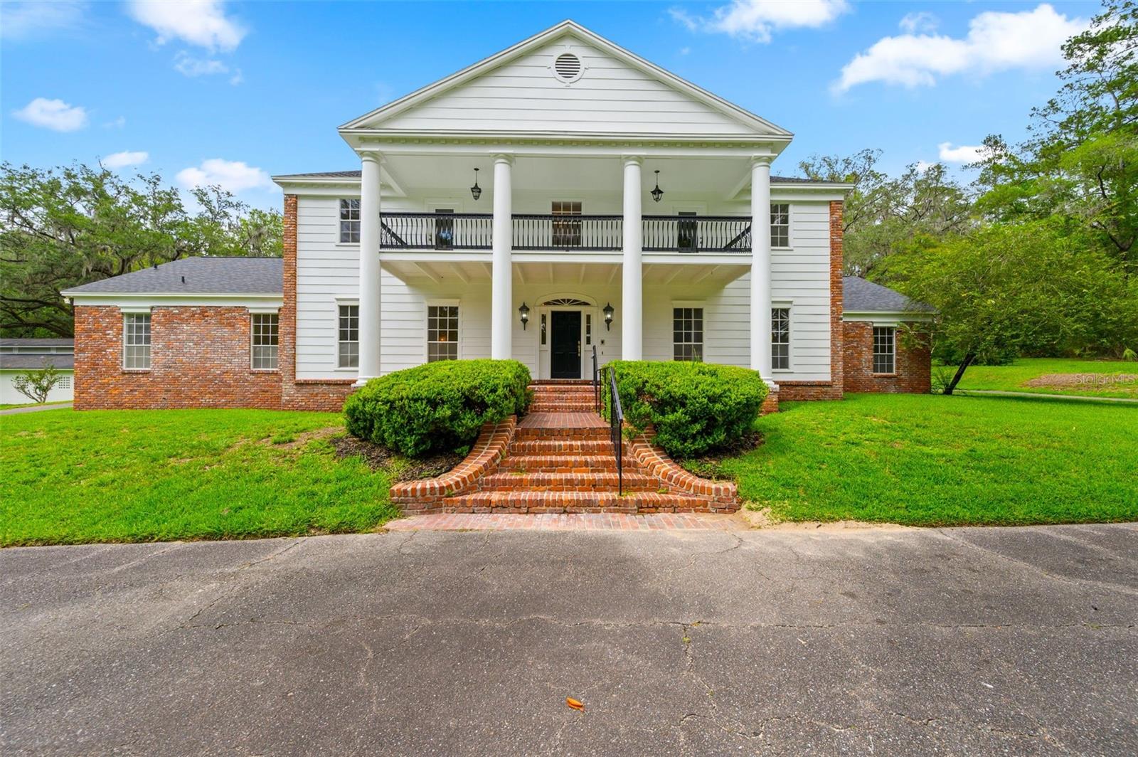Behind wrought iron gates and a brick fence on nearly 5 private acres, this magnificent colonial estate offers over 6,000 SF of luxury living in one of NW Gainesville's most exclusive — and best-kept — secrets. No HOA. No compromises. A circular driveway leads to a striking façade with tall round columns and dual balconies. Inside, nearly 20-foot foyer ceilings and a sweeping curved staircase set the tone. The traditional floor plan features a formal dining room, formal sitting room with fireplace, and a spacious kitchen with island flowing into a café area and family room anchored by a double-sided wood-burning fireplace. On the other side of that fireplace, a custom mahogany bar with built-in shelving, lighting, and sink makes the Parlor an entertainer's showpiece. The first-floor primary suite opens via French doors to the garden and pool lanai, with an adjoining study and guest half bath. A Florida sunroom with pool bath overlooks the large pool and travertine paver deck. Upstairs offers five oversized bedrooms and three full bathrooms. The property also features a large barn with true horse stalls and tack rooms — a rare find this close to everything. Minutes to Fresh Market, Thornebrook Village, Publix, Buchholz High, Santa Fe College, UF, UF Health, and Oaks Mall. A legacy estate of this scale, setting, and location is a once-in-a-market opportunity.