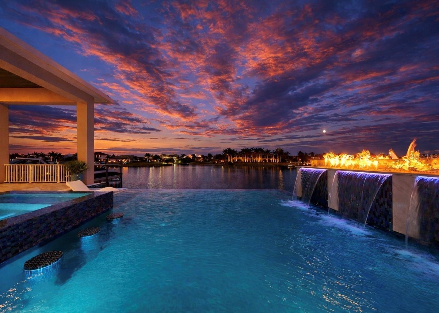 Exquisite waterfront home in Bay Estates Preserve at MiraBay offers resort-inspired coastal living at its finest. Set along a tranquil lagoon with direct access to Tampa Bay and the Gulf, this customized waterfront residence blends architectural precision and coastal design. Completed in 2022 and positioned on a .41-acre homesite with 100 feet of waterfront views, the home was designed to capture panoramic water vistas while offering the comfort of a single-story layout spanning 4,640 sq ft. A paver driveway, landscape lighting, cedar accents, and hurricane-impact glass entry doors introduce a residence where craftsmanship is immediately evident. Inside, fifteen-foot ceilings, crown molding, custom millwork, and oversized porcelain tile create open, elegant interior. WHOLE-HOUSE HURRICANE-IMPACT WINDOWS and expansive pocketing sliders blur the line between indoor and outdoor living, filling the home with natural light and water views. The main gathering areas flow seamlessly between the living room, dining space, and family room, where architectural details, designer lighting, and fireplaces create both sophisticated and welcoming spaces. A built-in bar with illuminated glass cabinetry enhances the home’s entertaining appeal. At the center of the home, the chef’s kitchen features an impressive eleven-foot quartz island beneath handcrafted wood ceiling beams. Monogram professional appliances include a six-burner gas rangetop, double ovens, Advantium speed oven, and integrated refrigeration. Bespoke cabinetry with integrated lighting and designer backsplashes complete the space. The owner’s suite offers a private waterfront retreat with lagoon views and automated window treatments. Entered through custom wood-and-glass barn doors, the spa-inspired bath includes a freestanding soaking tub, rain and aromatherapy shower, floor-to-ceiling tile, quartz vanities, and backlit mirrors. A customized boutique-style wardrobe room with handcrafted cabinetry and a quartz-topped center island adds exceptional luxury. Outdoor living defines the home. Beneath a 1,250-square-foot covered lanai with wood ceilings, coastal breezes flow across a spectacular infinity-edge pool and champagne spa finished in glass tile and surrounded by leathered marble decking. Waterfalls cascade along a privacy wall while a natural gas fire feature reflects across the water, creating an atmosphere perfect for both quiet evenings and gatherings. A sun shelf, swim-up seating, color-changing lighting, and pool automation enhance the experience. Entertaining continues in the outdoor culinary pavilion featuring a natural gas grill, stainless Kamado grill, and custom Gaucho grill, complemented by stone countertops, refrigeration, an ice maker, and illuminated storage overlooking the canal. For boating enthusiasts, a maintenance-free Trex dock extends over protected waters with a remote-controlled 16,000-pound lift, shore power, and water service, accommodating vessels up to thirty-five feet with direct access to Tampa Bay and the Gulf. Modern systems include dual Trane HVAC with UV purification, a tankless gas water heater, R-49 insulation, smart irrigation, whole-home water softening, structured wiring, Wi-Fi expansion, advanced security, and a three-car garage with EV charging. Residents of MiraBay enjoy resort-style amenities including waterfront clubhouse dining, heated pools, fitness facilities, tennis and pickleball courts, and kayaking. Schedule your private tour today.