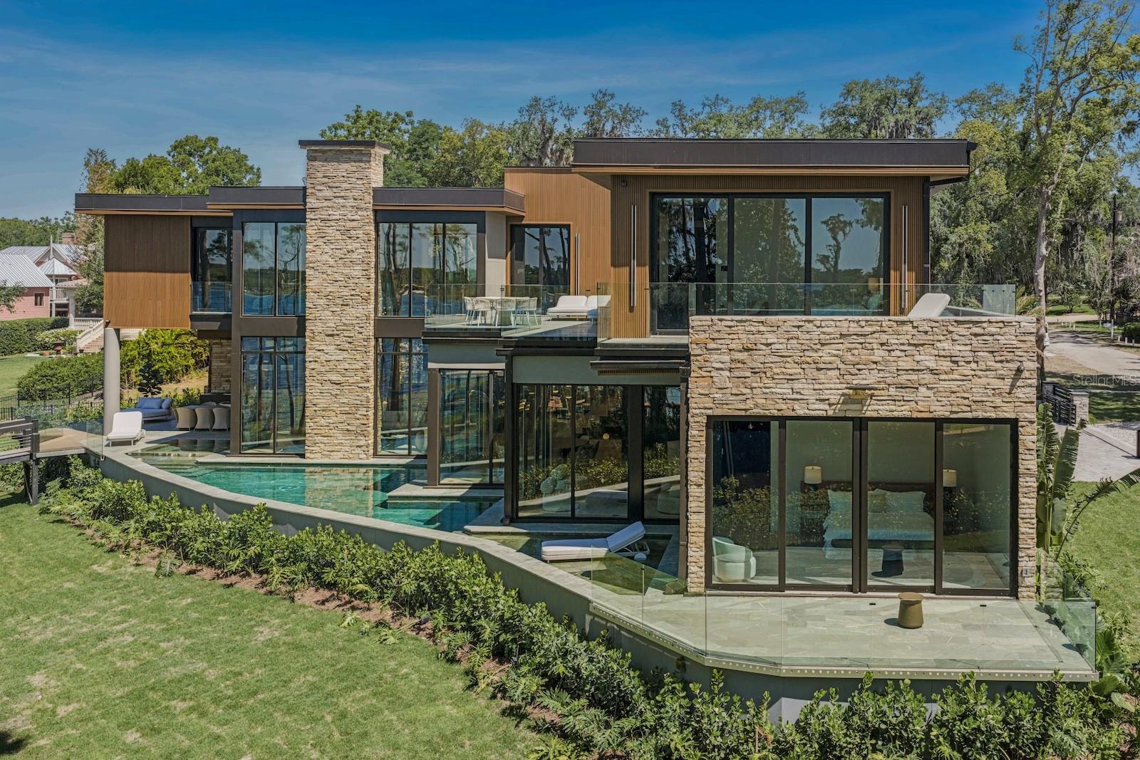 An extraordinary brand-new contemporary lakefront estate on prestigious LAKE BUTLER, part of the renowned Butler Chain of Lakes, offering an exceptional blend of architectural sophistication, modern luxury, and unparalleled waterfront living. Situated on approximately 0.83 acres with 114 feet of pristine lake frontage, this remarkable residence was designed to capture breathtaking panoramic water views, spectacular sunrises, and unforgettable sunsets from nearly every major living space. Encompassing 6,457 square feet of heated living area, the home seamlessly integrates architecture and landscape through expansive walls of glass, soaring ceilings, refined finishes, and an abundance of natural light. Every space was carefully oriented to maximize the connection between the interior and the surrounding waterfront setting. The designer chef’s kitchen showcases premium Sub-Zero and Wolf appliances, custom European cabinetry, an oversized waterfall island, Italian marble accents, and contemporary finishes, making it both a stunning focal point and an ideal space for entertaining. Designed for flexibility and comfort, the residence features six private bedroom suites, including two luxurious primary suites—one conveniently located on the main level and another on the second floor. Both primary retreats offer spa-inspired bathrooms with elevated finishes, expansive custom closets, premium TOTO fixtures, and living spaces that blend comfort and sophistication. The home also includes eight full bathrooms, one half bath, a dedicated wine room with elegant Italian marble detailing, wide-plank engineered hardwood flooring throughout, designer lighting, dual laundry rooms, a natural gas fireplace, and fully integrated Control4 smart home automation. Control4 home automation allows seamless control of lighting, audio, security cameras, pool equipment, and HVAC systems, creating a connected living experience. Outdoor living is equally impressive, featuring expansive covered lanais, a summer kitchen, saltwater pool and spa, and a private two-level covered dock equipped with water, electricity, and a boat lift. Designed for the ultimate waterfront lifestyle, the dock accommodates a 25-foot boat and two jet skis while providing direct access to the Butler Chain of Lakes, one of Central Florida’s most sought-after boating destinations. Constructed with exceptional attention to detail, the residence features concrete block construction on the main level, metal-frame construction on the second floor, DensGlass exterior sheathing for enhanced durability and weather resistance, energy-efficient TPO roofing, advanced spray foam insulation, four Daikin variable-speed HVAC systems with a combined 20-ton capacity, and natural gas tankless water heaters with recirculation technology. The garage is also equipped with dual 220V outlets for future EV charging capability. Located within the historic Town of Windermere, residents enjoy a unique golf-cart-friendly lifestyle with access to community parks, tennis courts, playgrounds, private lake amenities, and charming tree-lined streets, and the added benefit of no HOA, all while being just minutes from Orlando’s finest dining, shopping, world-class golf, and top-rated private schools. A rare offering on Lake Butler, where architectural excellence, luxury finishes, advanced technology, and one of Central Florida’s most coveted waterfront settings come together to create an exceptional living experience.