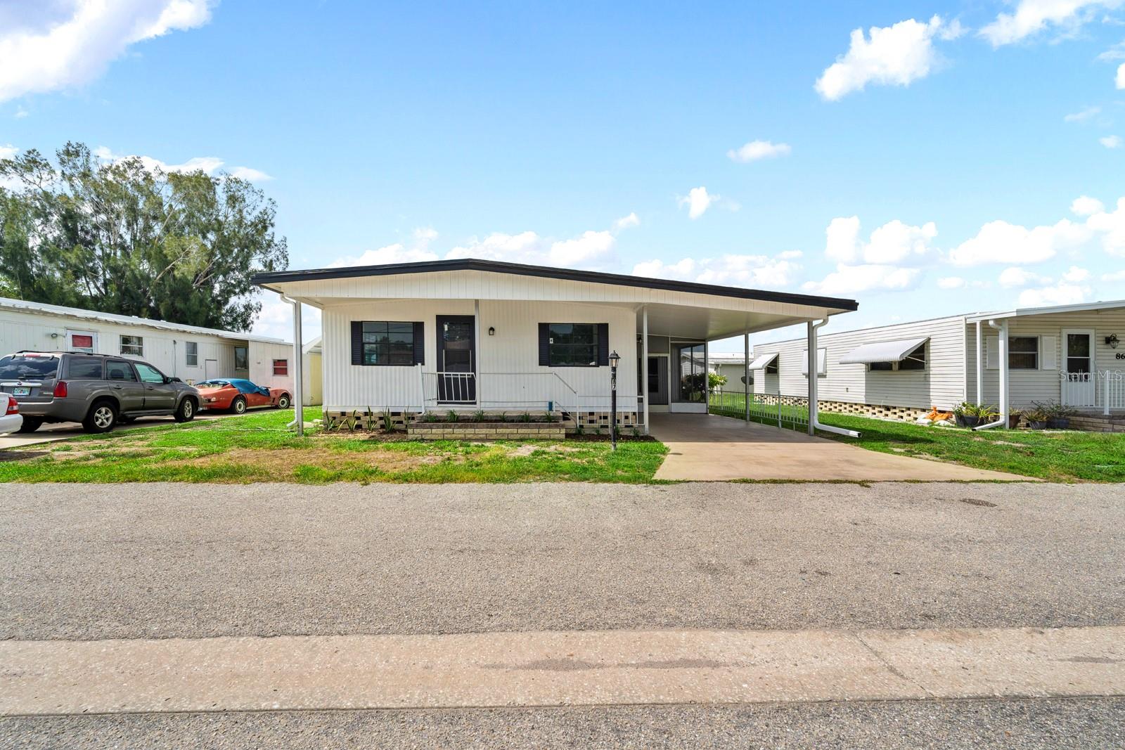 This inviting single-story manufactured home blends bright, modern interiors with comfortable everyday living. The open-concept living and kitchen area features abundant natural light, a soft neutral palette, wood-look flooring, white cabinetry with gold-tone hardware, stainless-steel appliances, and a breakfast bar with seating.

The home includes serene bedrooms and updated bathrooms, with details such as a marble-look tiled shower, contemporary fixtures, and warm metallic accents. An additional unfurnished room offers flexibility for a bedroom, home office, or hobby space. A dedicated utility area provides laundry connections, a utility sink, shelving, and a water heater.

Outside, the property offers a covered, screened porch, concrete driveway with parking, green lawn, and low-maintenance exterior. Aerial and street-level views show the home in an established manufactured-home community with neighboring residences, mature greenery, and convenient road access.

Ideally situated close to the clubhouse and heated pool, this non-age-restricted community allows up to two pets. Low quarterly fees include water, sewer, cable, trash collection, and internet, providing added convenience and value.