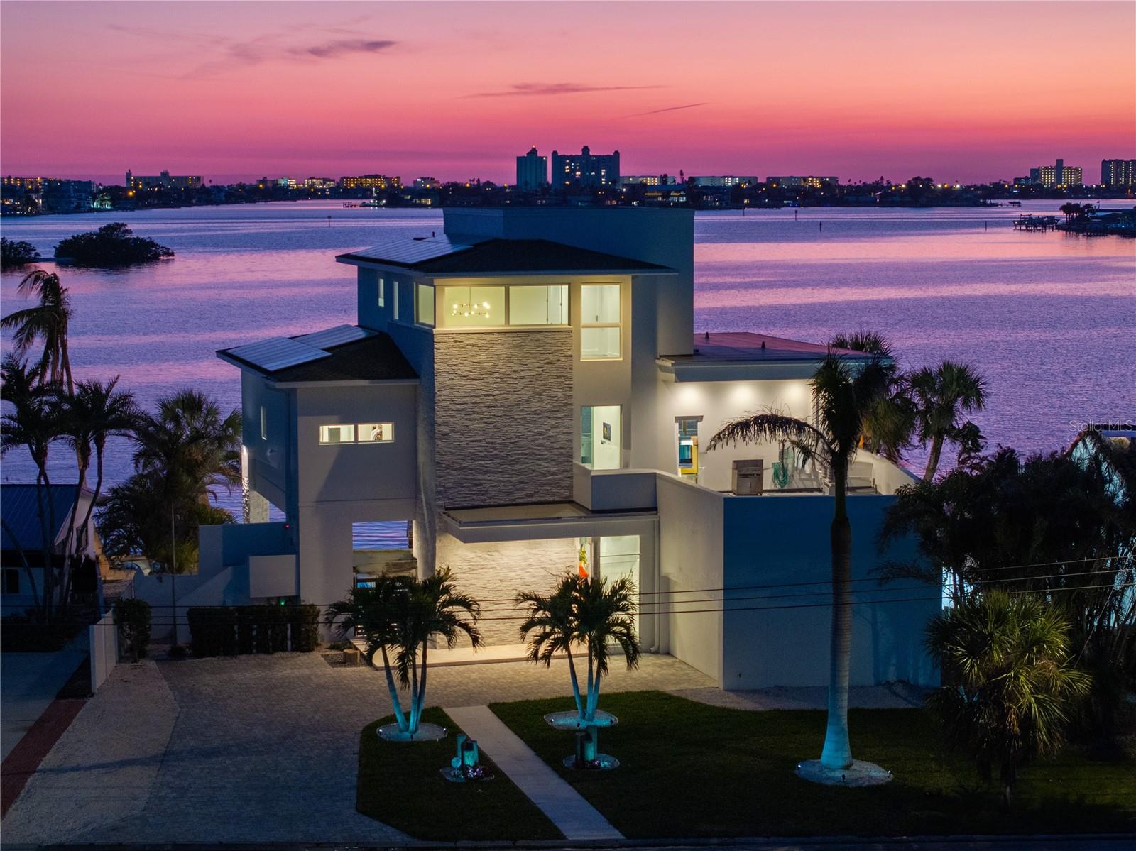Experience unparalleled coastal living in this custom-built, contemporary waterfront home, designed by Scott Geresy, architect and built by Tom Pici and Son. SITUATED 17 FEET ABOVE SEA LEVEL and NO WATER INTRUSION in the DWELLING. This exceptional residence offers breathtaking water views from every room and boasts top-tier construction, energy efficiency, and modern design elements. Built with superior craftsmanship, this home features block construction on two floors, supported by steel-reinforced concrete footings and cement block piers, ensuring unmatched structural integrity. The PGT WinGuard hurricane-rated windows/doors and breakaway walls provide exceptional storm protection, while the new exterior paint (2024), made from a salt-weather-resistant coating, enhances durability against the coastal elements. Designed for waterfront living, this home includes a newer seawall (2017), a large composite dock with a lower dock, swim ladder, and tie poles, as well as a 10,000 lb boat lift, making it ideal for boating enthusiasts. The saltwater, heated pool and hot tub, complete with a sunset shelf, create the perfect outdoor oasis, complemented by an expansive under-house shaded entertainment area and a rooftop terrace over the garage, offering breathtaking panoramic sunset views. Energy efficiency is a priority, with solar panels and spray/rigid insulation, significantly reducing electricity costs. A whole-house water softener and irrigation system ensure high water quality and low-maintenance landscaping. The home’s open floor plan with vaulted ceilings enhances its spacious feel, while floor-to-ceiling windows capture stunning water views from every angle. The chef-inspired kitchen features Silestone countertops, custom cabinetry, recessed lighting, pendant lighting, and top-of-the-line stainless steel appliances. Designed for comfort and convenience, the home is equipped with built-in blinds throughout and electric window shades in the dining and living areas. The huge custom closets by Southern Closets, including a laundry area and folding table in the primary suite, provide convenience and ample storage, while the guest bedroom features a secret second closet behind the built in shelves. The top-floor lounge includes a coffee area, perfect for relaxing with a morning cup while enjoying breathtaking views. Additional premium amenities include an ELEVATOR to all floors, an oversized garage with an electric car charger, and a dog run. The absolutely silent, energy-efficient A/C system is designed for easy access and maintenance, ensuring year-round comfort. A home security monitoring system provides added peace of mind. The combination of modern design, luxury features, and meticulous construction makes this residence a true waterfront gem. Nestled in a quiet and exclusive neighborhood with easy no bridge access, this contemporary masterpiece offers an extraordinary coastal lifestyle with unmatched durability, elegance, and breathtaking water views. Don’t miss the opportunity to own this dream waterfront home—schedule a private showing today!