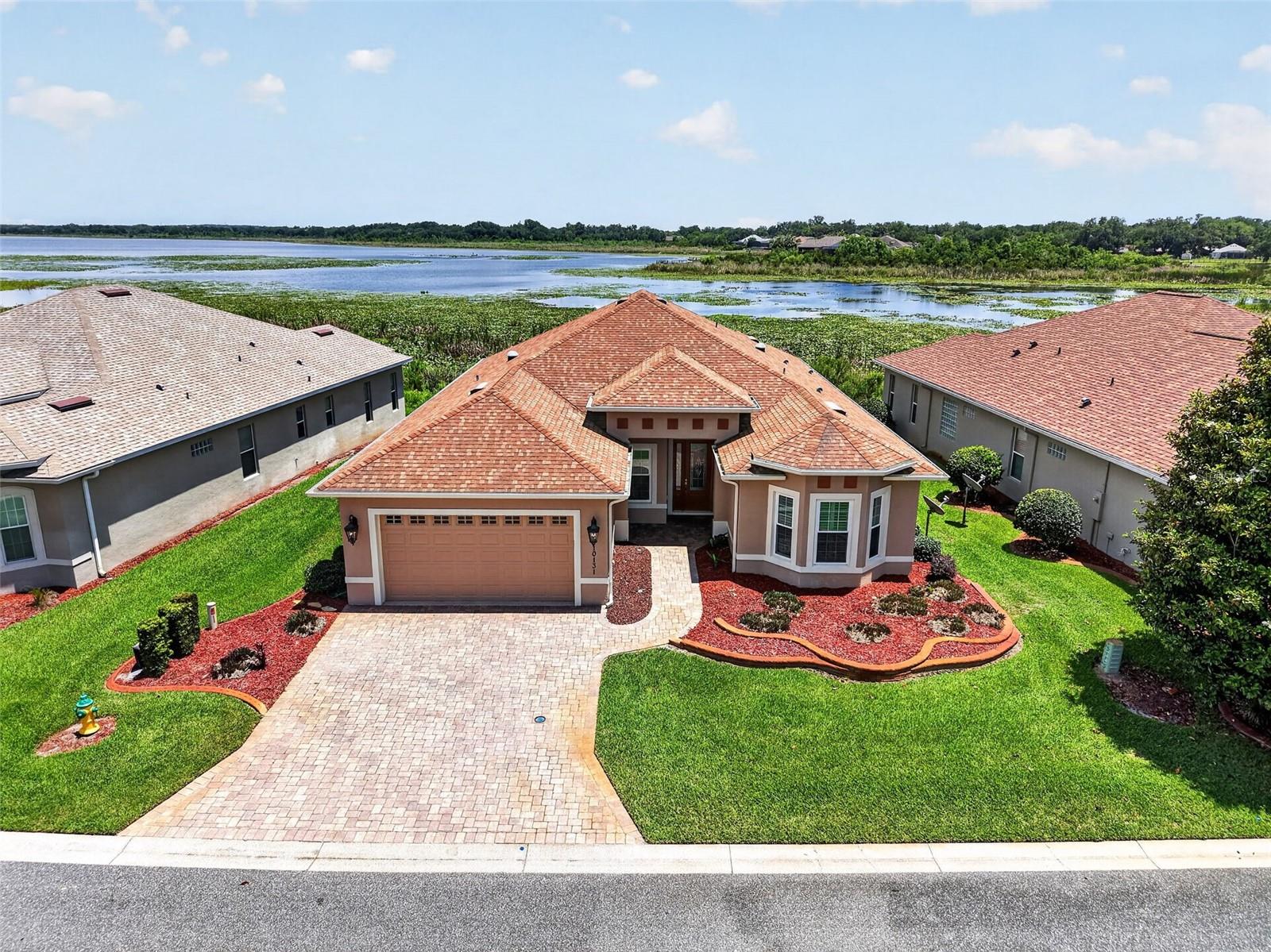Experience WATERFRONT LIVING at its finest in this beautifully maintained “LAKESIDE” model nestled within the GATED community of LAKESIDE LANDINGS. Offering 3 bedrooms, 2 baths, a den, and an expansive GLASS-ENCLOSED lanai, this exceptional home showcases BREATHTAKING panoramic VIEWS of LAKE MIONA and a lifestyle designed for comfort, beauty, and relaxation. From the moment you arrive, the inviting LEADED GLASS front door with sidelite sets the tone for the details found throughout. Inside, you’ll appreciate the seamless blend of ceramic TILE flooring in the main living areas and stylish LAMINATE flooring in all bedrooms — truly a NO CARPET home. The thoughtfully designed kitchen features GRANITE countertops, light cherry cabinetry with convenient upper and lower pull-out drawers in bottom cabinets, STAINLESS appliances including a SMOOTH TOP double oven range, French door refrigerator, microwave, and dishwasher. Just off the kitchen, the spacious laundry room offers PLANTATION SHUTTERS, abundant cabinetry, a built-in sink, and an oversized PANTRY for exceptional storage. FRENCH DOORS lead to the versatile DEN, complete with plantation shutters, making it an ideal OFFICE hobby room, or optional 4th bedroom. The spacious living room opens through sliding glass doors with elegant LUMINETTE SHEERS to the show stopping enclosed lanai — undoubtedly the HEART OF THE HOME — where tranquil LAKE VIEWS and abundant WILDLIFE create a peaceful retreat you’ll enjoy year-round. The PRIMARY SUITE is a true sanctuary with LAMINATE flooring, TRAY ceiling, large BAY WINDOW overlooking the lake, and two WALK-IN closets leading to the EN-SUITE BATH featuring GRANITE countertops, dual sinks, custom framed mirrors, a Roman WALK-IN SHOWER, and private water closet. A pocket door creates a PRIVATE GUEST WING with two generously sized guest bedrooms offering LAMINATE flooring, PLANTATION SHUTTERS, ceiling fans, bay window, and ample closet space. The guest bath includes GRANITE countertops and a separate tub/shower area with glass enclosure. Additional highlights include a painted garage floor, INSULATED garage doors, paver driveway and walkways, decorative curbing, gutters surrounding the home, and maintenance-free lawn care provided by the HOA — allowing you more time to enjoy the incredible amenities and lifestyle this community offers. Residents of Lakeside Landings enjoy resort-style amenities including a SPECTACULAR POOL COMPLEX, FITNESS center, RECREATION CENTER, library, BILLARDS, TENNIS and PICKLEBALL courts, organized activities, social events, and more — all conveniently located adjacent to THE VILLAGES.