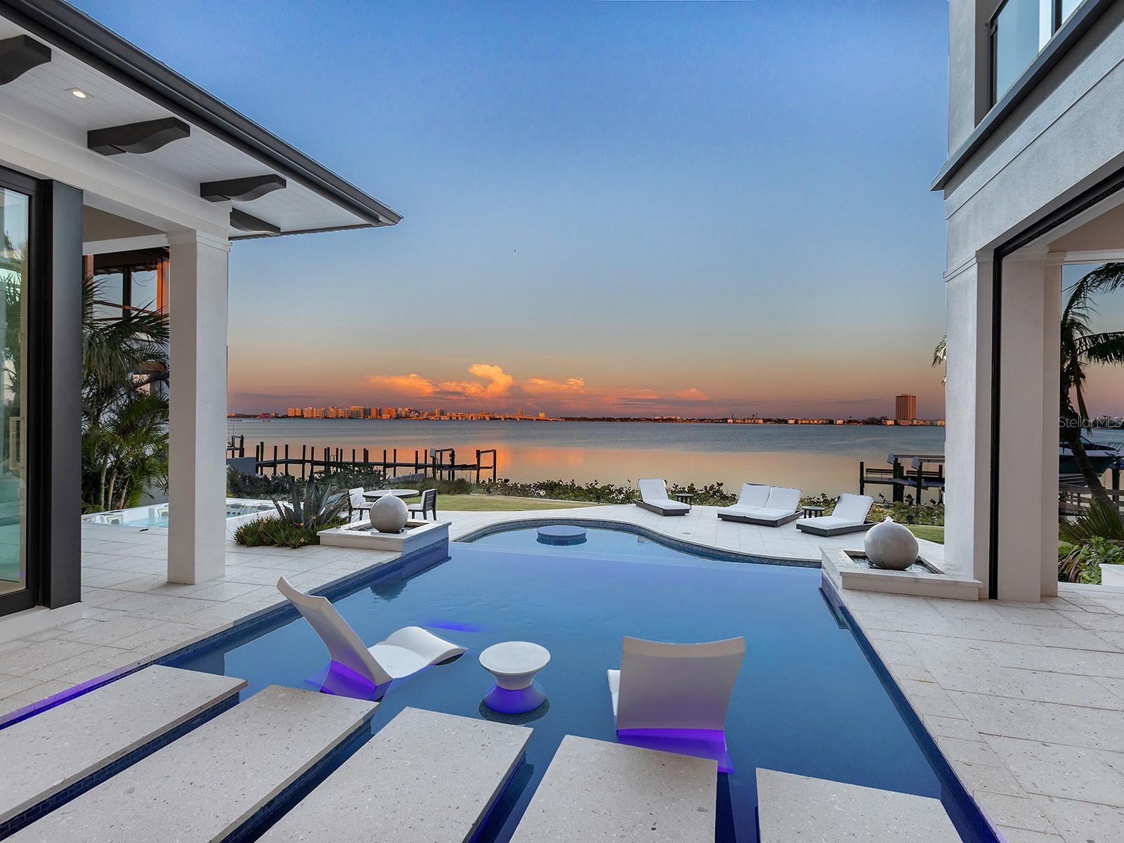 Under contract-accepting backup offers. Set against Sarasota’s coveted bayfront in Lido Shores, this extraordinary waterfront estate captures sweeping views of Sarasota Bay, the downtown skyline, Ringling Bridge, and City Island, offering an unparalleled luxury lifestyle with effortless access to boating, beaches, and downtown Sarasota. More than a residence, this is a statement property—an iconic example of West Indies–inspired architecture executed with exceptional craftsmanship and modern sophistication. A private gated entry introduces a home where over $1 million in enhancements and more than 2,000 hours of custom design are evident throughout. Advanced smart home automation allows seamless global control, blending innovation with refined living. The custom front door opens to a dramatic foyer and a great room defined by a coffered ceiling, media wall, linear fireplace, and expansive bay views. The dining space features a quartz-topped bar with wine storage and flows into a chef’s kitchen appointed with Thermador appliances, a large floating island with quartz waterfall edges, and a walk-in pantry with an additional Bosch refrigerator. The primary suite is a tranquil sanctuary with floor-to-ceiling glass, sliders, and breathtaking water views. Two custom walk-in closets complement a spa-inspired bath featuring floating vanities, a soaking tub, a custom multi-head shower, and sliders leading to a private Zen garden with an outdoor shower. A sculptural staircase and glass-enclosed wine wall lead to an upper-level loft with a wet bar and waterfront terrace. A refined office with custom built-ins and four en-suite guest bedrooms, each with custom closets, projector screens, and private balconies, ensure comfort and privacy throughout. Outdoors, the estate transforms into a resort-style waterfront retreat. An infinity-edge pool with sun shelf, in-pool table, and cascading spa anchors the setting, surrounded by multiple seating areas, a firepit, and an outdoor shower. The fully equipped outdoor kitchen—featuring a smoker, grill, wok, and side burner—makes alfresco entertaining effortless, while a spiral staircase leads to an upper terrace with panoramic views. The newer deepwater dock is designed for serious boating, offering a 20,000-pound boat lift, jet ski lift, water and power, ambient lighting, and a dedicated fishing area. With direct access to Sarasota Bay just steps from the backyard, this property delivers exceptional boating and waterfront living. Additional features include a three-car garage, two laundry rooms, an elevator, a garage drop zone, and a hidden utility closet. The home is fully integrated with Control4 automation, security cameras, a 48-kilowatt generator, 20-kilowatt battery backup, high-end audio-visual systems, automated shades, circadian lighting, outdoor theater, app-controlled pool and irrigation, Wi-Fi-enabled appliances, EV charger prewiring, Starlink backup internet, motion-activated lighting, and voice control via Josh AI. Lido Shores is one of Sarasota’s most sought-after and architecturally significant waterfront communities, offering residents exclusive access to the north end of Lido Beach and close proximity to St. Armands Circle, Longboat Key, City Island, and downtown Sarasota. This remarkable bayfront estate is an architectural triumph, designed for sophisticated entertaining, advanced living, and extraordinary waterfront luxury.