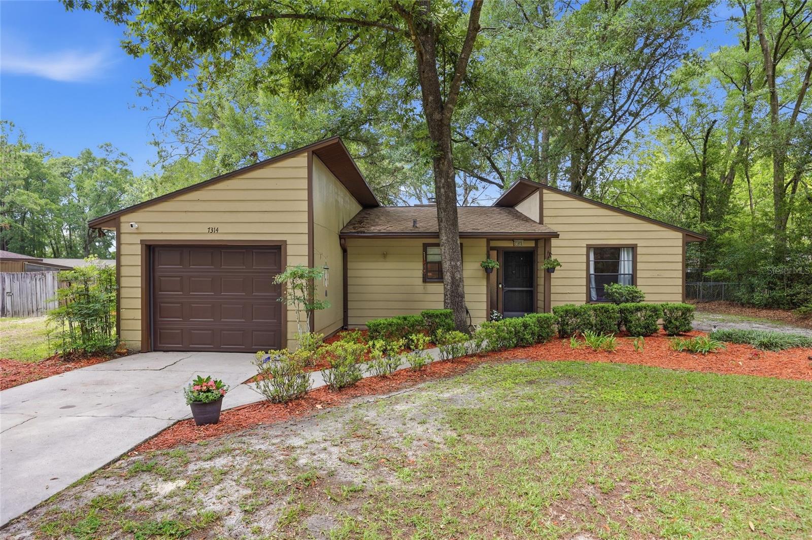 Shaded by a canopy of mature trees, this unique 1,299 sq ft, 3-bed, 2-bath contemporary home sits on a 0.23-acre lot, offering an architectural layout that stands out from the crowd! Boasting striking asymmetrical rooflines and a classic mid-century-inspired aesthetic, the property greets you with pristine landscaping and instant curb appeal. Step indoors and cross the threshold into a deep, tiled foyer equipped with a convenient closet. The layout flows gracefully into an expansive living and dining room where plush carpet underfoot leads your eyes to the warmth of a wood-burning fireplace. Adjacent to the main living space, the kitchen caters to the home chef with upgraded granite countertops, a beautiful backsplash, and plenty of cabinet space. This smart, split-bedroom plan ensures ultimate privacy for everyone. The primary suite showcases soft carpet, a large walk-in closet, and a tub/shower combination, while the 2 guest bedrooms also feature carpet and share an easily accessible guest bath updated with a modern walk-in shower. Located off the kitchen is the 1-car garage with a new garage door and opener. The garage houses the built-in laundry hookups for maximum convenience. Stepping through the kitchen’s back door, you will discover a fully fenced backyard complete with a patio that is ready for quiet mornings or weekend gatherings. Situated in the peaceful Pine Glade neighborhood with no HOA fees, this prime location sits right off Tower Road, just minutes from the premier shopping, dining, and entertainment options at Butler Plaza and Celebration Pointe. Daily errands are a breeze with a Publix-anchored shopping center right around the corner. A convenient 5-minute stroll takes you straight to the nearest Regional Transit System (RTS) bus stop on Tower Road, which can greatly simplify your daily commute. For leisure and outdoor recreation, the stunning Kanapaha Botanical Gardens and quiet local parks are located just down the road, while the University of Florida campus and UF Health Shands remain an effortless drive away. Don’t miss your chance to own this captivating property; schedule your private tour today!