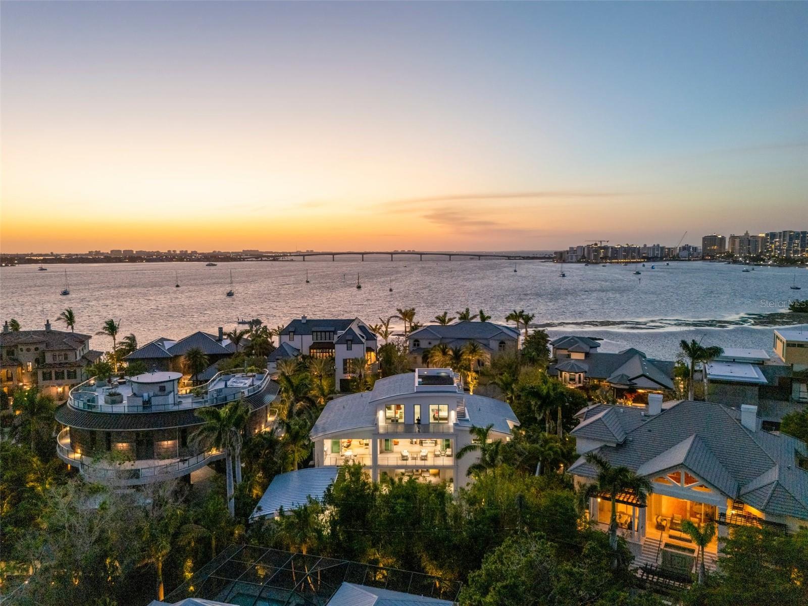 A striking triumph of modern architecture and engineering, this three-story contemporary estate in Harbor Acres—Sarasota’s most coveted neighborhood—stands as a bold statement of design, innovation, and uncompromising luxury. Designed by renowned architect Mark Sultana of DSDG Associates, the residence is a masterwork where precision and artistry converge. From the moment of entry, soaring ceilings, expansive walls of glass, and a dramatic Italian floating staircase set the tone. Natural light fills the interiors, enhancing a layout designed for both impact and livability. The main level is anchored by an open-concept great room and a chef’s kitchen of exceptional caliber. A full suite of Miele appliances—including induction cooktop, built-in espresso maker, warming drawers, dual microwaves, and a full-height wine tower—is paired with Dekton countertops and custom Westwood cabinetry. A concealed butler’s pantry with Sub-Zero refrigeration enhances functionality. Living and dining areas flow seamlessly to a covered terrace with a summer kitchen and thermally modified wood ceiling overlooking the private grounds. The north wing is dedicated to a dual-owner’s suite—two private retreats connected by a spa bath with floating vanities, dual rain showers, soaking tub, heated towel racks, and custom Italian closets. One suite is currently configured as a refined office. The second living level offers a library/media room that can convert to a two-room guest suite, an additional bedroom suite, and two full baths for comfort and privacy. Outdoors, a resort-style setting unfolds with a dramatic 50-foot heated pool and spa, covered lounge areas, and lush bamboo for privacy. A separate one-bedroom guest house with full bath, coffee bar, and mini refrigerator provides independent accommodations. The rooftop terrace captures sweeping panoramic views of Sarasota Bay, the Ringling Bridge, Longboat Key, Siesta Key, and the downtown skyline—an exceptional setting for sunset gatherings. Additional features include Crestron-controlled lighting, shades, and audio, integrated speakers, five Luma security cameras with remote access, and two climate-controlled garages for up to six vehicles with epoxy floors, storage, and a gym opening to the pool terrace. A rare architectural statement offering elevated design, privacy, and location just minutes from downtown Sarasota and the Bayfront.