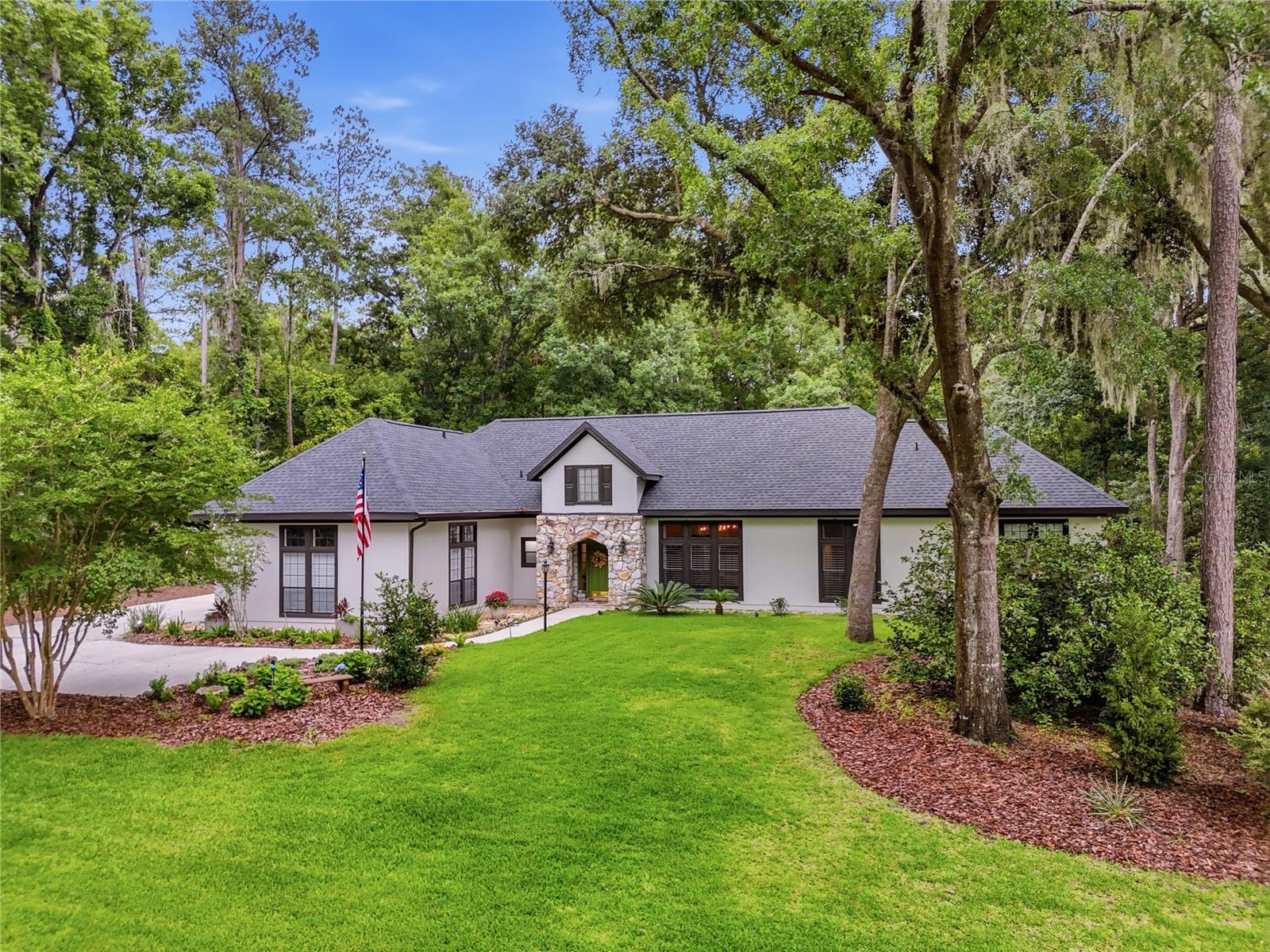 Set on a wooded 1.6-acre corner lot in Pine Hills Estates, this architect-designed Tudor home offers more than 3,477 square feet, extensive renovations, and a degree of privacy that is difficult to find in West Gainesville. The location provides the space and seclusion of a larger homesite while remaining just a short drive from the shopping, dining, and everyday conveniences of Town of Tioga.

Originally built as the architect’s personal residence, the home features oversized rooms, abundant natural light, and architectural details rarely found in newer construction. The layout includes three bedrooms, two and a half bathrooms, two separate living areas, a formal dining room, and a private office that could also serve as a fourth bedroom, guest room, den, or hobby space.

The renovated kitchen includes custom floor-to-ceiling cabinetry, quartz countertops and backsplash, an oversized island with seating and storage, a professional-style gas range, double wall ovens, a farmhouse sink, glass-front display cabinetry, and a large walk-in pantry. It opens directly to the main living area, where a floor-to-ceiling stone fireplace, custom built-ins, and a wall of windows overlook the backyard.

A second living room near the front of the home includes another fireplace, large windows, and direct access to the enclosed Florida room. The formal dining room connects to the Florida room through French doors, allowing the home’s main gathering spaces to flow together while maintaining clearly defined rooms.

The spacious primary bedroom includes a gas fireplace, large windows overlooking the wooded grounds, access to the Florida room, and custom California Closet systems. The primary bathroom has been fully renovated with a long dual vanity, custom cabinetry, illuminated mirrors, extensive storage, and a large glass-enclosed shower with designer tilework. The secondary full bathroom has also been completely renovated, and the half bathroom has been updated.

Additional details throughout the home include crown molding, transom windows, skylights, French doors, custom built-ins, stone accents, plantation shutters, and three gas fireplaces.

Multiple sets of French doors open to an expansive enclosed Florida room with walls of windows, skylights, ceiling fans, and space for both dining and sitting areas. From there, the home opens to a large screened lanai with paver flooring and plenty of room for outdoor dining and entertaining. An adjoining open patio overlooks the wooded backyard.

Mature hardwoods, established landscaping, garden paths, stone borders, and a wooded buffer provide separation from neighboring homes while leaving usable lawn and garden areas around the house.

The property also includes an attached two-car garage, a detached third-car garage that could serve as a workshop or hobby space, and a separate storage building toward the rear of the lot. The broad driveway provides generous parking and easy access to both garage areas.

This is an ideal West Gainesville home for buyers seeking updated interiors, flexible living space, substantial indoor and outdoor entertaining areas, and a private setting within easy reach of Town of Tioga and the rest of Gainesville.