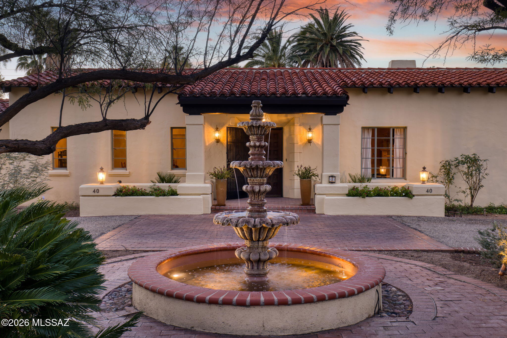 Historic elegance abounds at 40 E Plaza del Encanto, set on nearly an acre within Tucson's prestigious El Encanto Estates and offering a rare opportunity to own a piece of the city's architectural history. This exquisite estate blends timeless craftsmanship with serene desert living and urban sophistication. Built in 1934, this Spanish Colonial Revival home was one of the original El Encanto homes, with design overseen by renowned architect Lewis ''Starky'' Starkweather. Behind the gated north semi-circle driveway, handcrafted wrought-iron fencing, mud adobe walls, mature trees, and a grand fountain set a tone of privacy and sophisticated charm. The stunning handcrafted mahogany door opens into an inviting foyer with double-arched entries that showcase dramatic scale and (continued) architectural character, including a grand living room and an elegant formal dining room designed for distinctive gatherings, whether entertaining or family.  Soaring ceilings, crown moldings, fireplace with wood mantle and marble threshold, distinctive chandeliers, and gorgeous, original oak floors (which extend throughout the residence), are prominent features. The sun room, with gorgeous terracotta cement tile, extends along the south side of the home, featuring expansive picture windows and a full-length covered brick patio to reinforce indoor/outdoor living. This room seamlessly connects the east kitchen and the west-side library room, both of which feature mahogany floors. The updated kitchen, with an adjacent butler's room, is both elegant and functional, featuring a chef-grade Viking range, two sinks, wood-and-glass-front cabinetry, a generous peninsula with pendant lighting, vintage-style fixtures, and a large, bright picture window framing the lush desert landscaping beyond. To the south of the kitchen, a large ensuite area features a private bedroom, guest bathroom, and a sitting room with double French doors opening onto a covered patio overlooking a grass lawn and a sparkling pool area. Directly across the lawn, the home's west wing includes three large bedrooms, each with a private bath and oak flooring throughout. The primary suite features a sitting area with a gorgeous fireplace, a separate dressing area with double closets, and a tiled bathroom. Double French doors open onto an east-facing covered patio, with the south patio, lawn, covered ramada, fire pit, and pool area beyond. Within the walled backyard area, an independent heated/cooled space with a wood ceiling and exposed brick walls offers an opportunity for an office, music room, or art studio. Recent upgrades include replacement of all gas lines, newer heating and cooling units for the main house, complete built-up roof recoatings (garage, studio, house), pool replaster, tiling, and equipment upgrades, fountain replaster/upgrades, and the addition of an automatic solar-powered wrought iron driveway gate on the east side of the property. Lovingly restored, this home exudes historic authenticity, beauty, and peace in Tucson's most exclusive central neighborhood, a true desert sanctuary within urban vibrance. After more than 22 years off the market, this storied residence awaits its next steward to experience Tucson's heritage at its most refined.