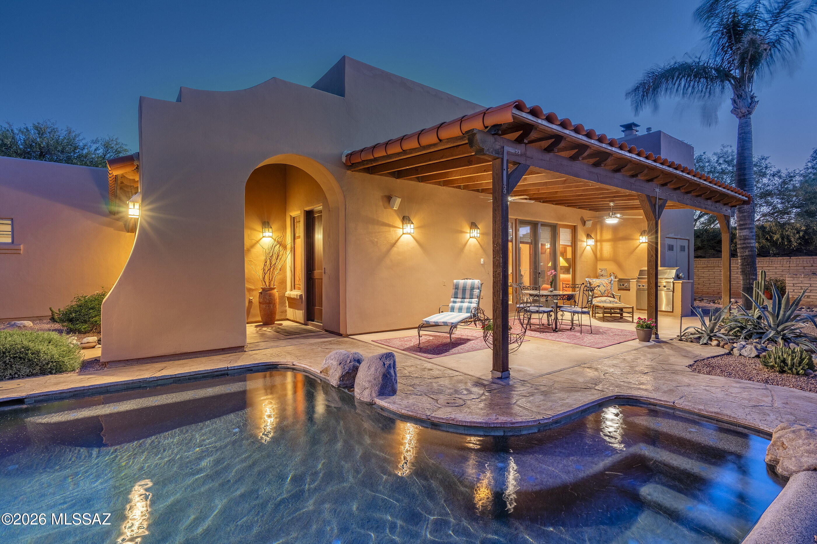 Situated within the gated enclave of The Garrison in the Old Fort Lowell Historic District, this residence offers a level of architectural presence and design integrity that is both rare and immediately felt. A graceful arched entry introduces a private, walled courtyard, setting a tone of understated sophistication and intentional privacy from the moment of arrival. Inside, the home unfolds with a striking sense of volume and light. Soaring ceilings, articulated by exposed wood vigas, create a dramatic vertical scale, while clerestory windows are positioned with precision, allowing natural light to enter as a dynamic element within the space. Throughout the day, light shifts and softens, subtly transforming the interiors and shaping the overall atmosphere. The result is an environment that feels expansive yet grounded, where openness and light influence the experience of the home in a way that is both calming and quietly compelling. Stained concrete floors anchor the space, complemented by softly contoured walls and a refined, cohesive material palette.

The Great Room serves as the architectural and social center of the home, where living, dining, and kitchen spaces flow together seamlessly. Expansive glass doors and large format windows frame views into the surrounding courtyard, dissolving the boundary between indoors and out while amplifying both scale and livability.
The kitchen is cleanly integrated yet distinctly appointed, featuring rich cherry wood cabinetry carried throughout the residence, paired with slab granite countertops and a full height granite backsplash. A professional grade stainless steel range and hood create a strong focal point, while the center island with bar seating extends naturally into the living space, offering both function and connection.

The primary suite is privately positioned and generously scaled, with direct access to the courtyard and a soft infusion of natural light. The ensuite bath reflects a continuation of the home's material language, with dual vanities, a soaking tub set within a tiled surround, and a glass enclosed shower with detailed tile work, all executed with restraint and consistency. Secondary spaces offer flexibility and thoughtful separation, each maintaining a connection to light and outdoor access.

At the center of the home, an all encompassing courtyard wraps around three sides of the residence, serving as both the visual anchor and the defining lifestyle element. A pool with a natural rock water feature introduces movement and texture, while covered patios, exposed beams, and multiple seating areas create a series of outdoor rooms designed for both gathering and retreat. Masonry walls and curated desert landscaping provide a sense of enclosure and privacy, while multiple points of access from within the home reinforce a seamless indoor outdoor experience.