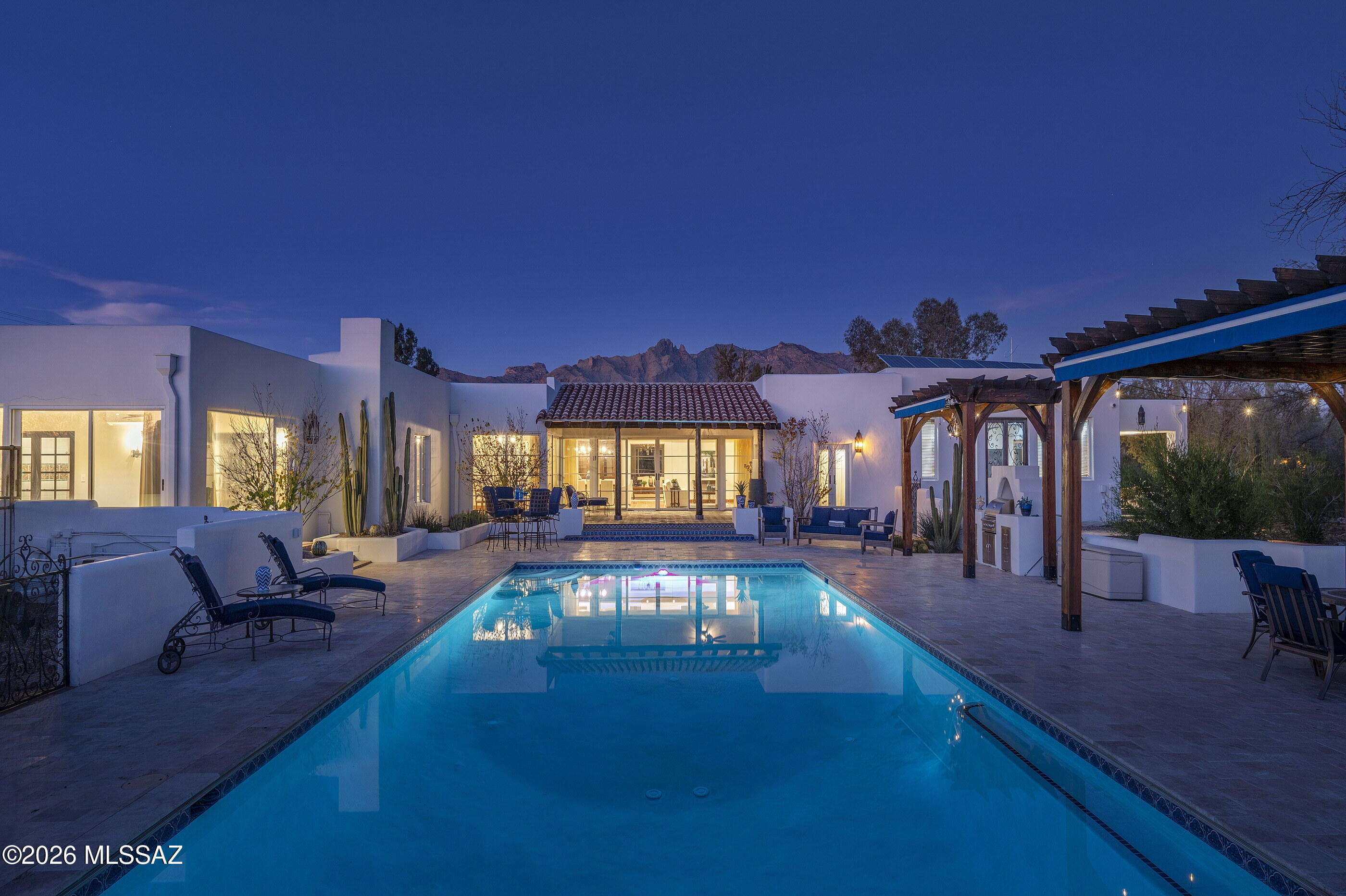 Behind a private gate and situated on over six acres, this estate stands as a testament to Josias Joesler's architectural imagination. Thick adobe walls, hand-formed and enduring, rise against the desert sky, while clay tile roofs and smooth white plaster walls give the home a presence that is both timeless and grounded. The landscape feels inseparable from the architecture: courtyards, patios, and terraces are carefully composed to catch light, frame views of the Catalina Mountains, and draw the desert inside, where every room has a sense of connection and flow. Walking through the grounds, there is a feeling of quiet discovery, as if the house reveals itself gradually, its history embedded in the weight and texture of its walls. Inside, the estate is defined by material richness, with hand-troweled plaster and exposed wood beams introducing warmth, texture, and rhythm, while custom millwork and artisan-crafted details articulate the hand of the maker. Terracotta tile flooring stretches through the principal rooms, grounding the interior in an organic dialogue with the desert beyond. Decorative tiles, subtly placed, punctuate hallways, kitchens, and baths, offering moments of craftsmanship and tactility that hint at the house's romantic revival lineage. Light is treated as a design element: carefully positioned picture windows and walls of glass frame the mountains and courtyard, drawing in the changing moods of the day and allowing interiors to shift from intimate sanctuary to luminous gathering spaces.

The kitchen is both functional and expressive, a space that feels naturally central. A generous island of polished stone, streaked with striking blue-green veins, commands attention without overwhelming, while custom wood cabinetry balances warmth and refinement. Glass-front display units provide lightness, and pendant lights in aged metal and glass introduce moments of rhythm and presence, echoing the lines of the exposed beams above. Here, cooking, conversation, and quiet reflection coexist seamlessly; the room feels alive yet grounded, a space built for everyday life as much as for entertaining.

Living and dining areas continue this interplay of openness and intimacy. Interiors flow directly to patios, covered porches, and outdoor rooms, blurring the boundaries between inside and out. Each transition is deliberate, each threshold designed to frame a view, offer shade, or create a pause. The pool sits within a broad natural stone terrace, providing ample room for lounging and dining, while pergolas with substantial wood posts, built-in seating, and outdoor kitchens establish zones for gathering. Desert-adapted plantings, mature trees, and careful landscaping create privacy and serenity, making the grounds feel simultaneously expansive and protected.

The estate feels like a world of its own, a desert sanctuary where space and tranquility define every moment. Yet this sense of seclusion is perfectly balanced: within minutes, the city's most vibrant experiences are at your doorstep. La Encantada's boutique shopping and fine dining, midtown's cafés and galleries, and downtown Tucson's cultural heartbeat are all just moments away. Here, the rare intimacy of private desert living coexists effortlessly with immediate access to the city's best amenities, creating a balance that transforms everyday life into something both serene and effortlessly connected.
