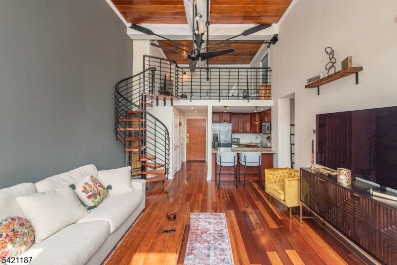 Volume, light, and character come together in a way that feels visually integrated and distinctly different from anything else at The Mill. This one bedroom residence is defined by its dramatic double-height living space, where soaring ceilings, exposed brick, and a full wall of oversized windows create a setting that is both open and striking. Natural light pours across rich hardwood floors, highlighting the scale of the room and the cohesive layout. Overhead, dual ceiling fans and track lighting accent the height of the space, while the brick backdrop and deep window sills bring in that signature Mill aesthetic. Positioned just off the living space, the kitchen is finished with warm wood cabinetry, granite countertops, stainless steel appliances, and a breakfast bar that offers seating. A custom full-sized loft sets this unit apart. Accessed by a eye-catching spiral staircase, the loft adds a completely separate level of living, ideal for a lounge, office, or creative space. Framed by industrial-style railings and set beneath wood-accented ceilings with exposed steel elements, it overlooks the main level. The bedroom continues the same clean aesthetic with hardwood flooring, oversized windows and complemented by a sliding barn-style closet system that adds design. This is a one-of-a-kind layout within The Mill, is rarely found.